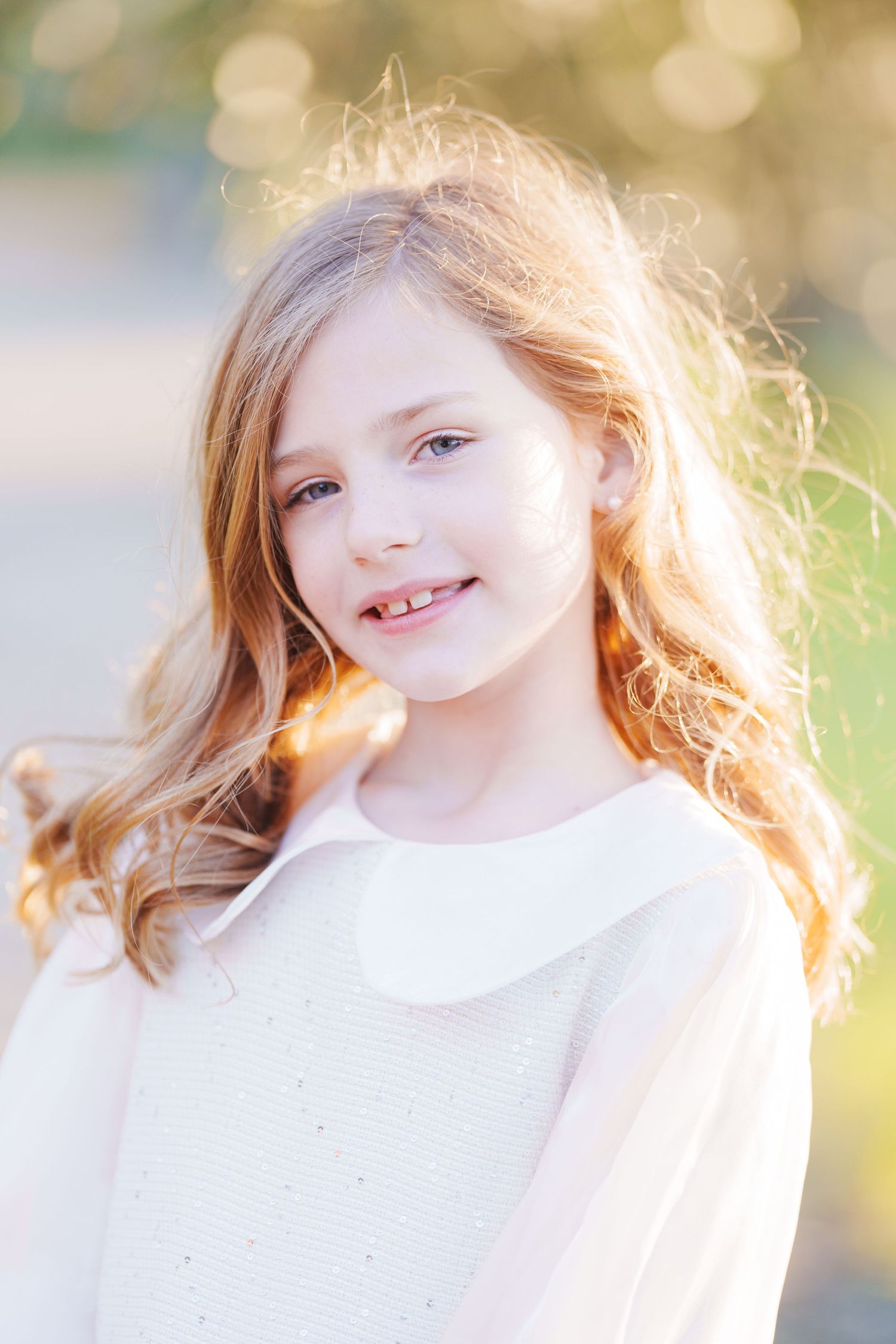 Smiling young girl with long blonde hair wearing a white dress outdoors in soft natural light.