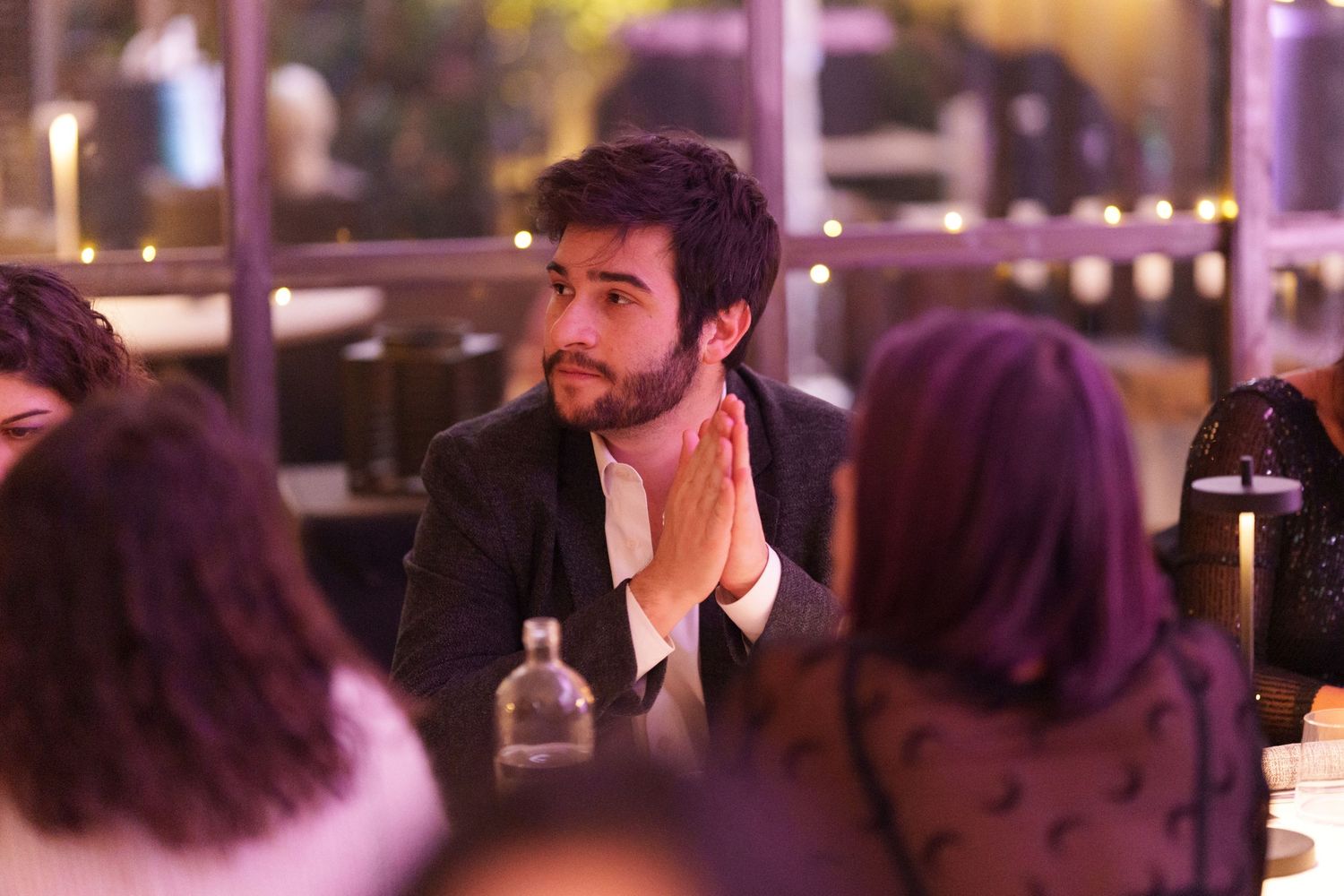 Man with beard in dark blazer sits at dinner table with hands clasped, surrounded by guests in warm ambient lighting.