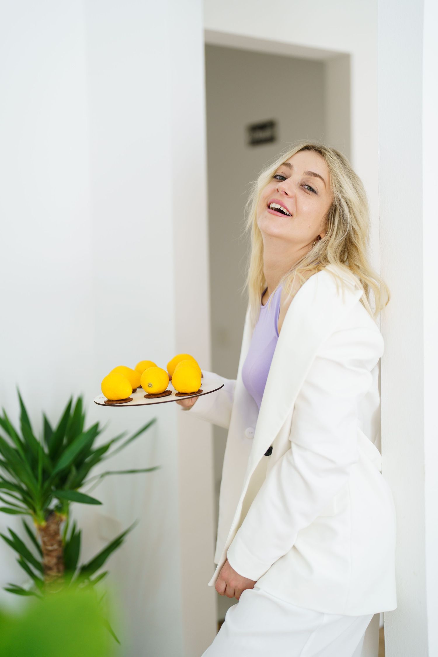 Smiling blonde woman in white suit holding a plate of fresh yellow lemons in a bright modern interior.