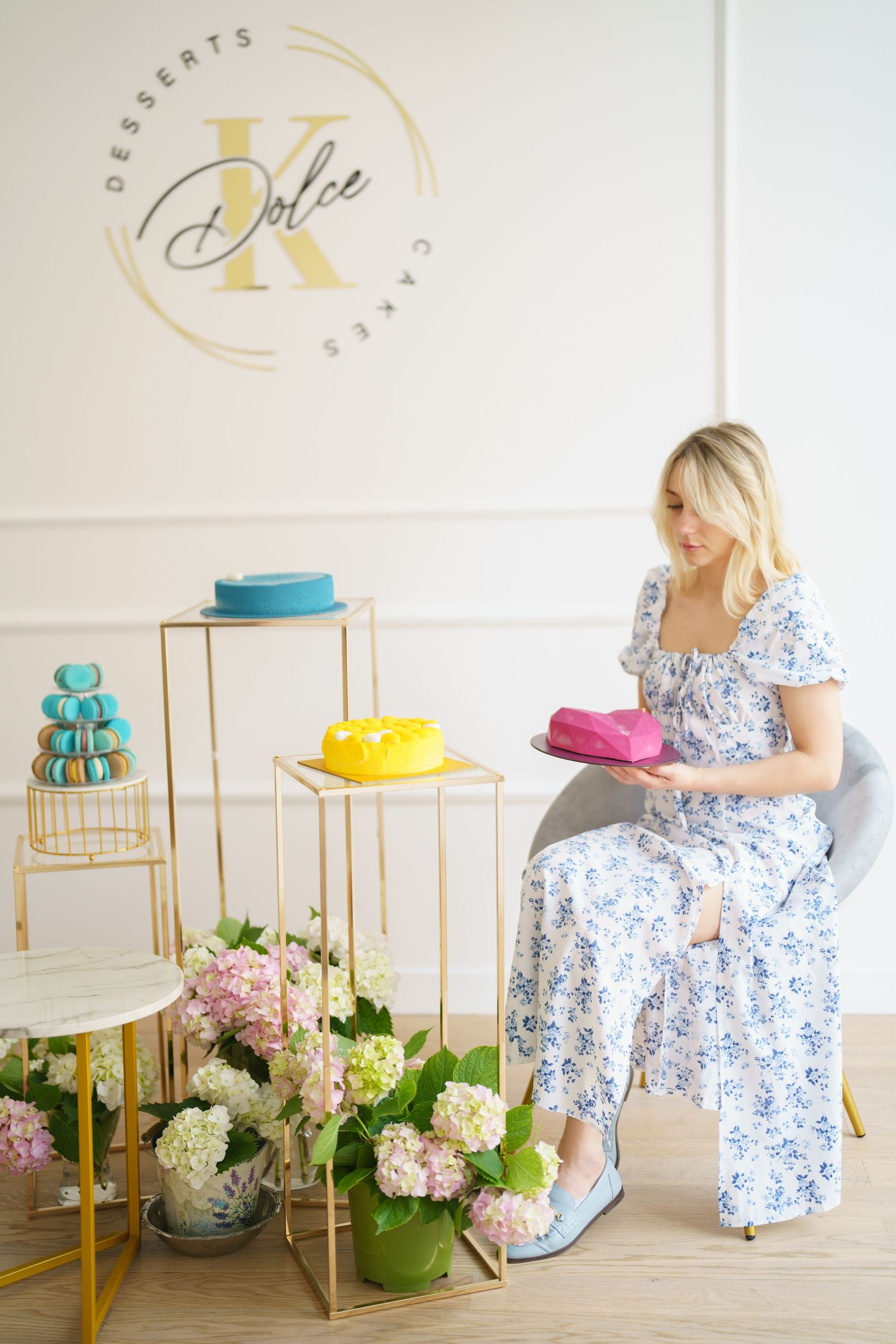 Blonde woman in floral dress holding colorful cakes at Dolce K dessert shop with macarons and flowers.