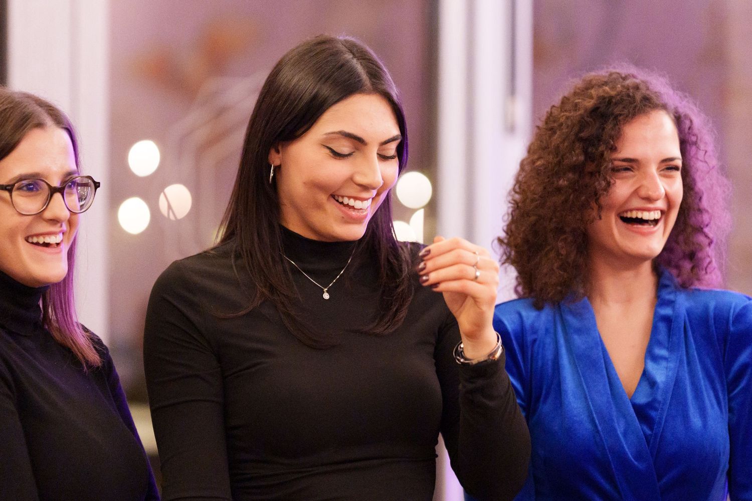 Three women smiling and laughing together at an indoor event, wearing elegant black and blue outfits.