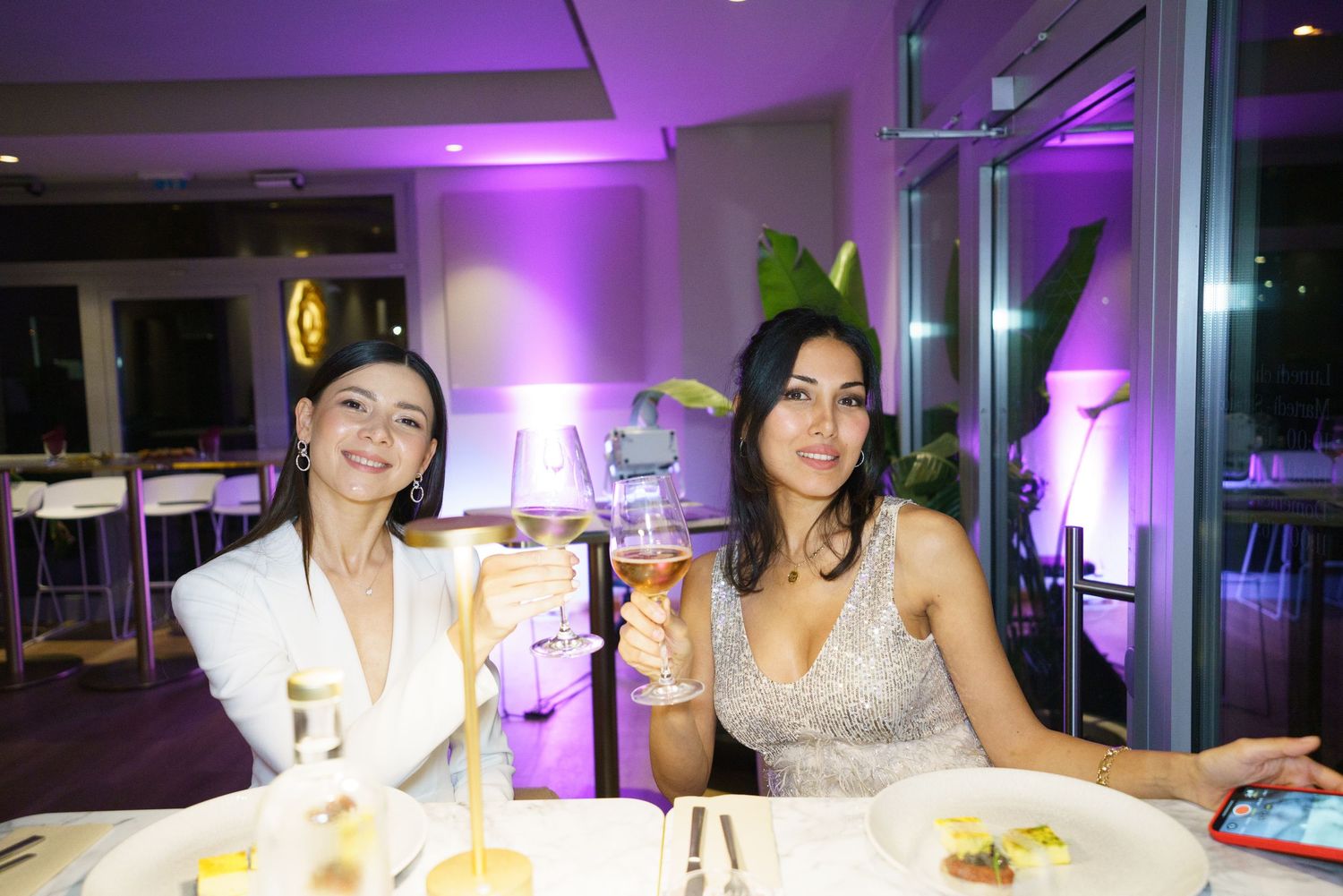 Two women smiling and toasting with cocktails at an elegant restaurant with purple ambient lighting.