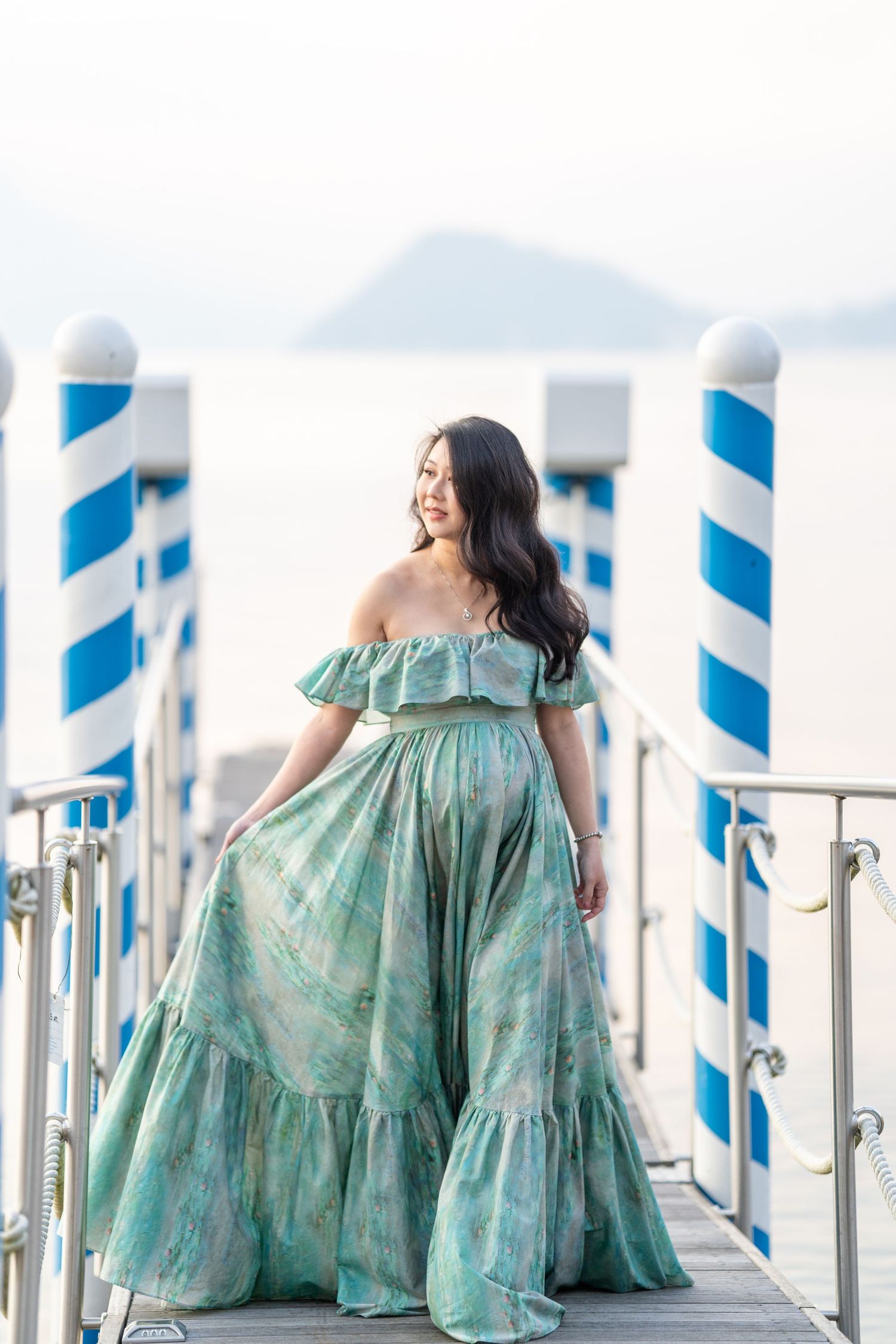 Woman in green off-shoulder floral maxi dress poses on a seaside pier with blue and white striped poles.