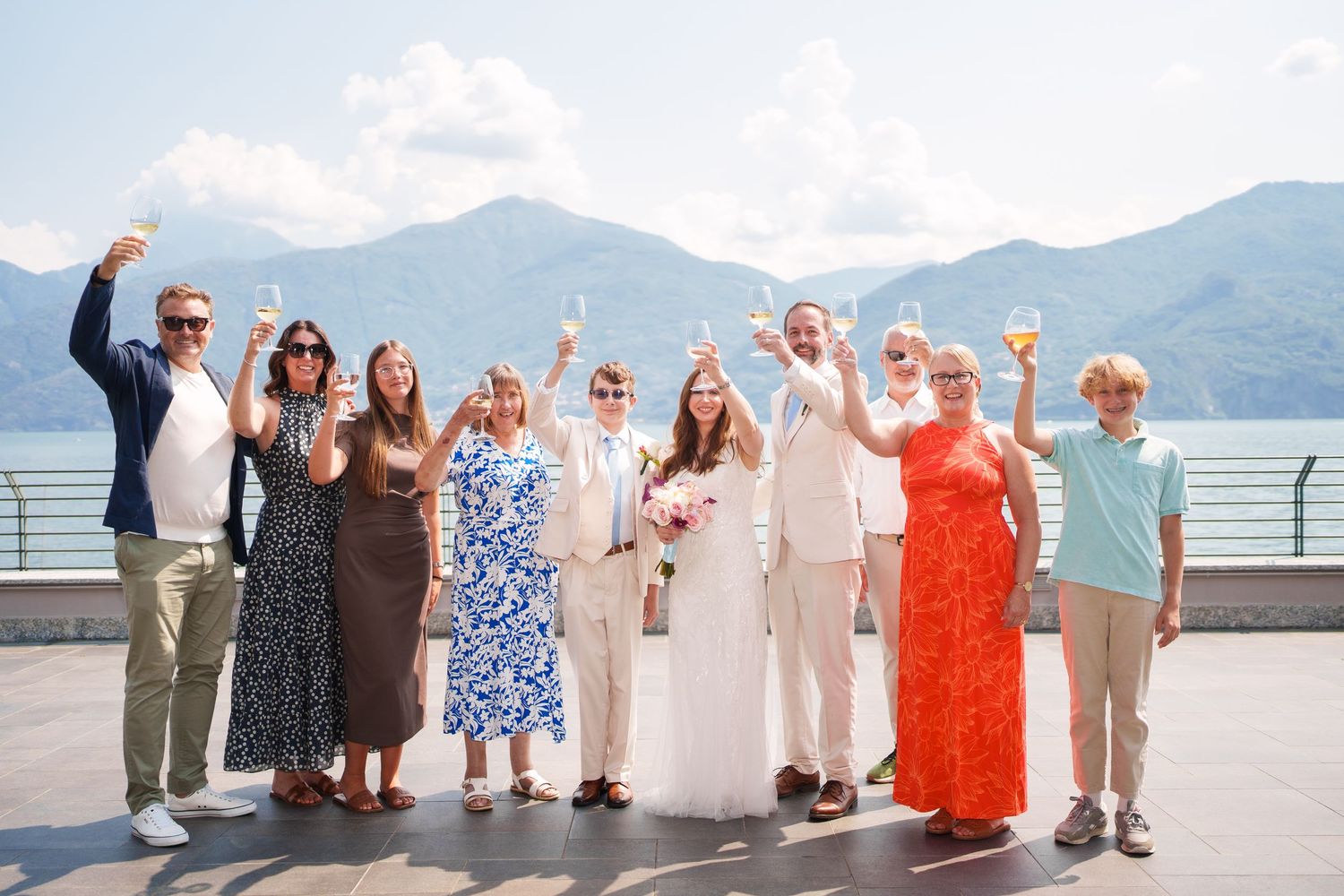 Wedding party toasting with champagne on a lakeside terrace with mountain views on a sunny day.
