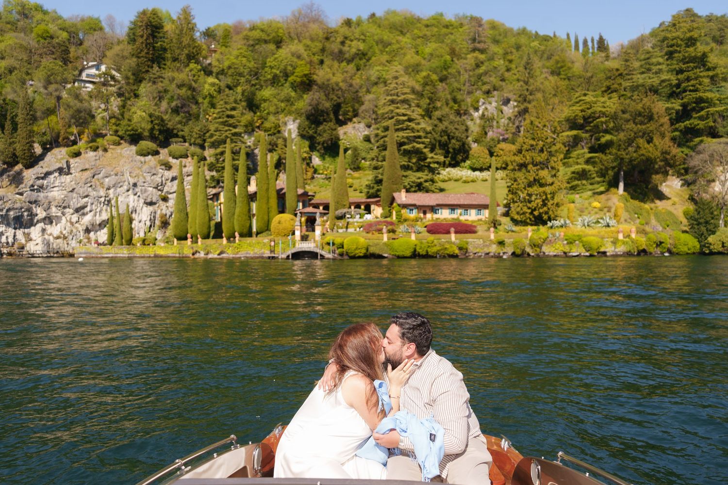 Couple kissing on a wooden boat on a lake with lush green hillside villa in the background.