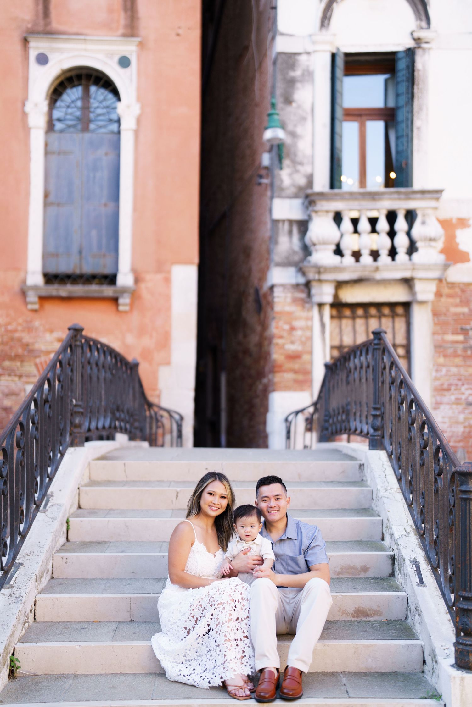 Family of three sitting on stone bridge steps in Venice, Italy, with historic architecture in the background.