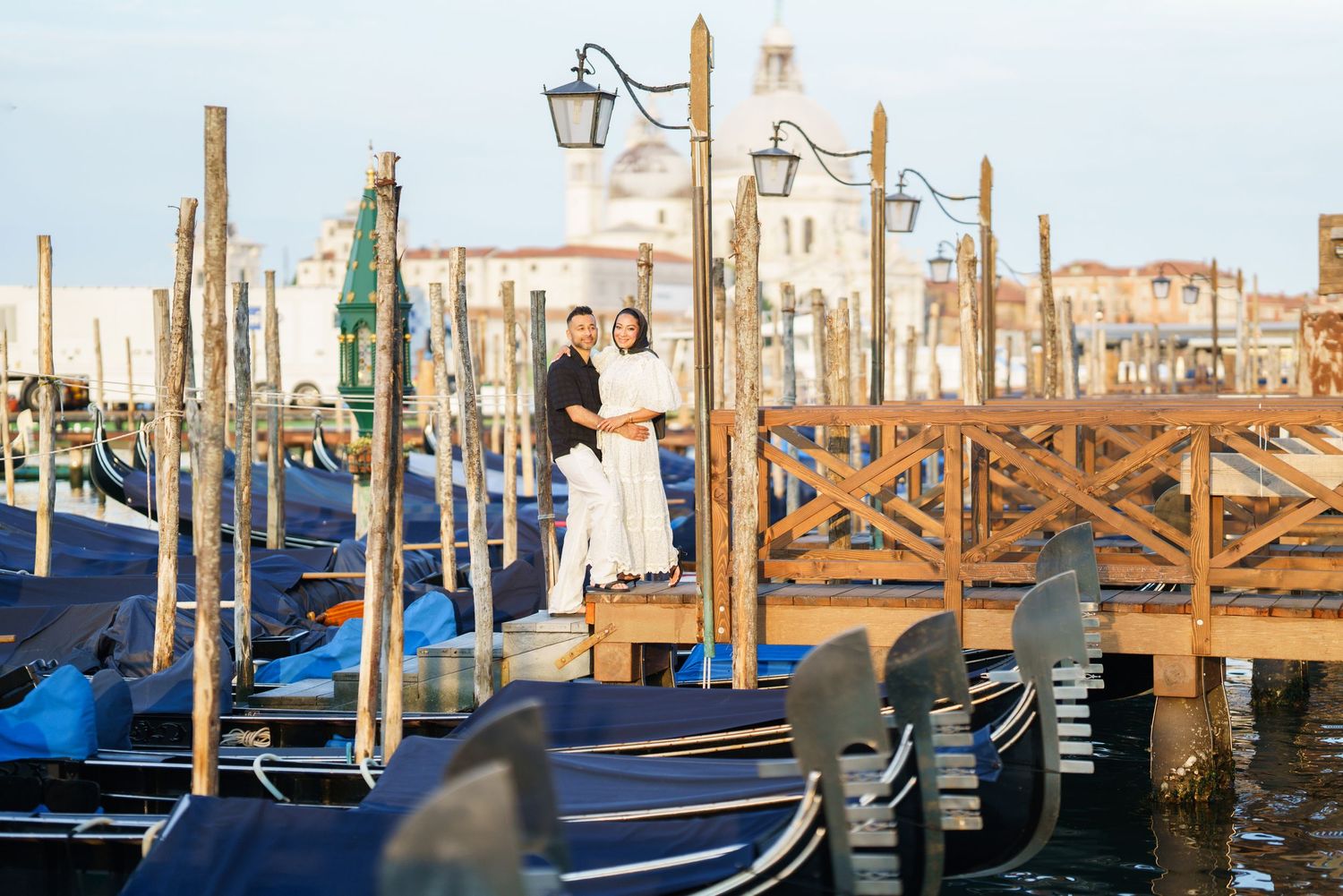 Couple embracing on a wooden dock surrounded by gondolas in Venice, Italy with historic buildings in background.