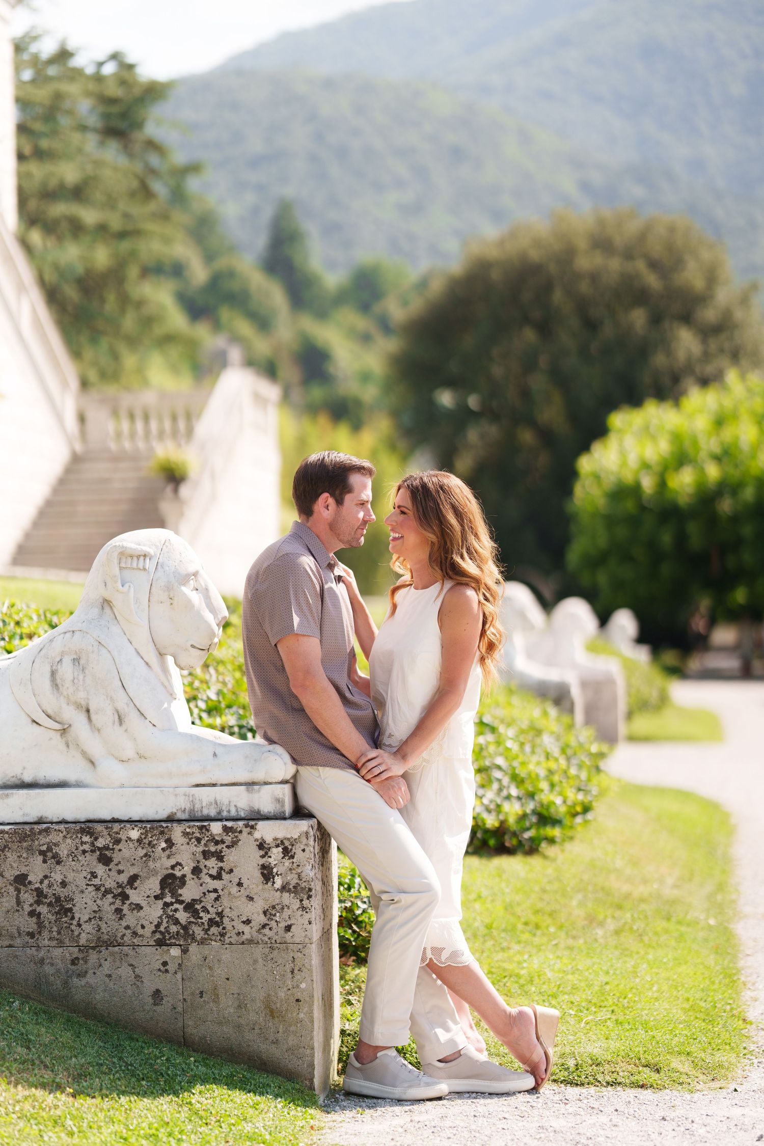 Couple smiling at each other in a lush garden with lion statues and mountain backdrop on a sunny day.