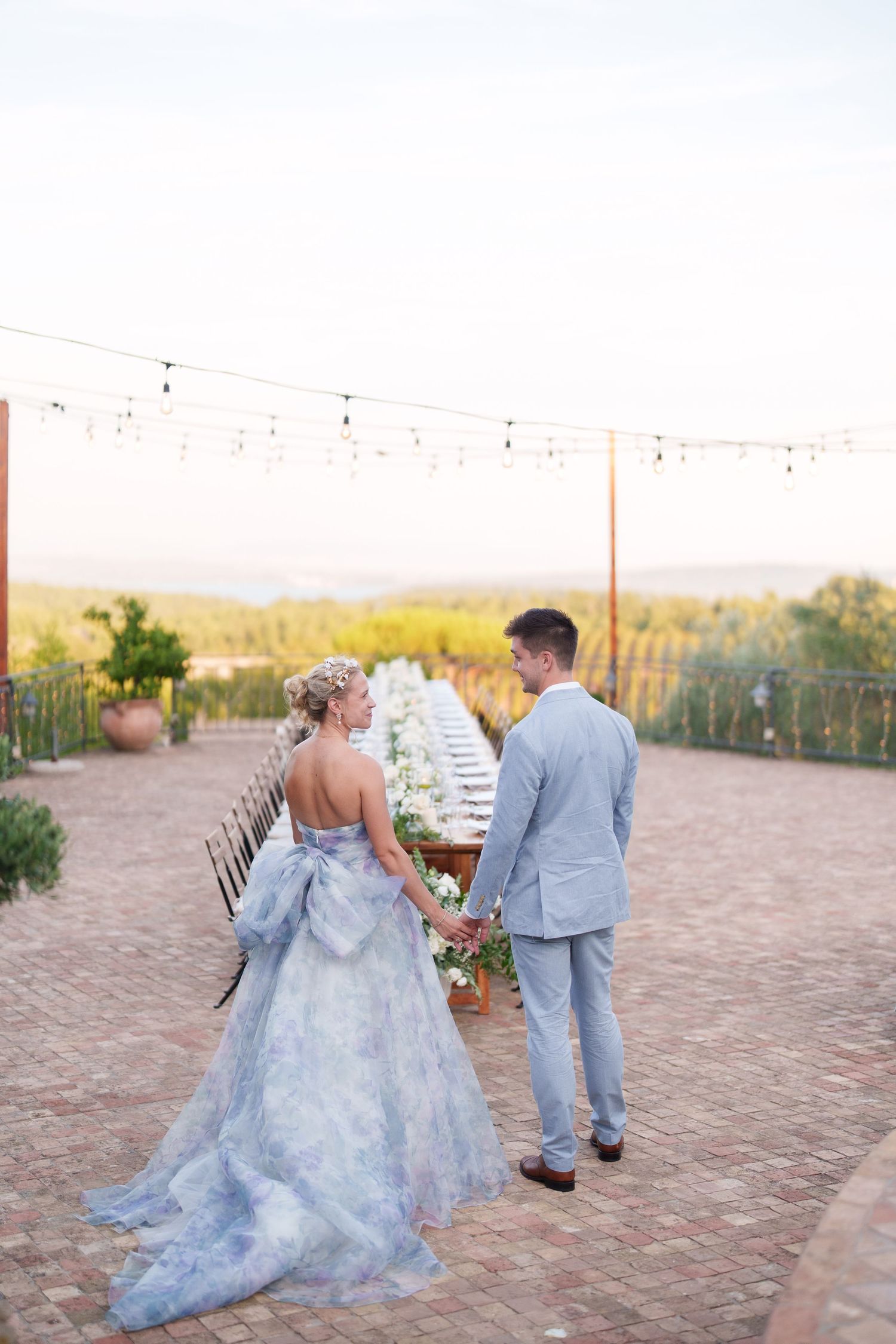 Bride in blue floral gown and groom in gray suit hold hands beside elegant outdoor reception table at sunset.