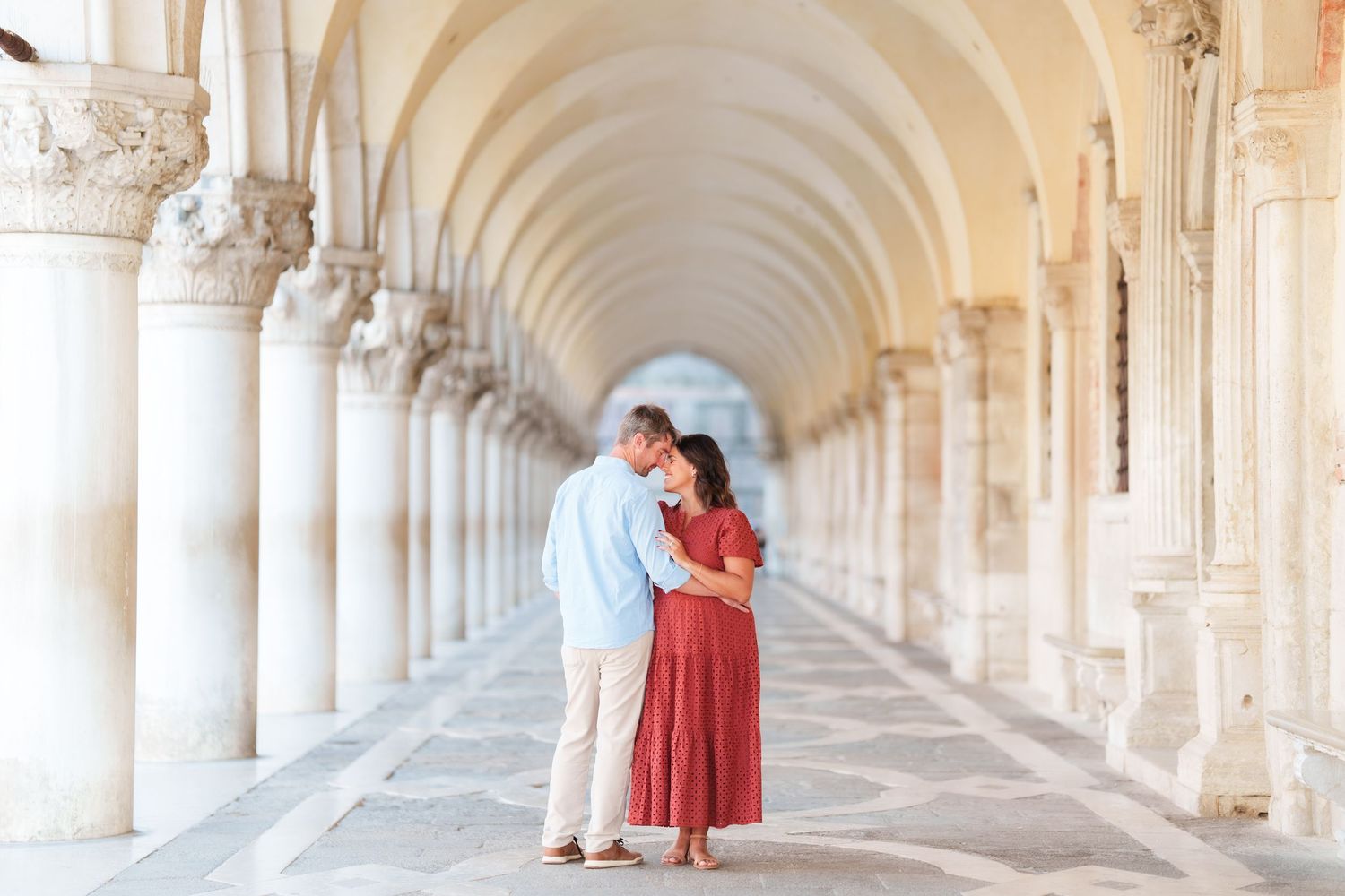 Couple embracing romantically under arched colonnade in Venice, woman in red dress, man in light blue shirt.
