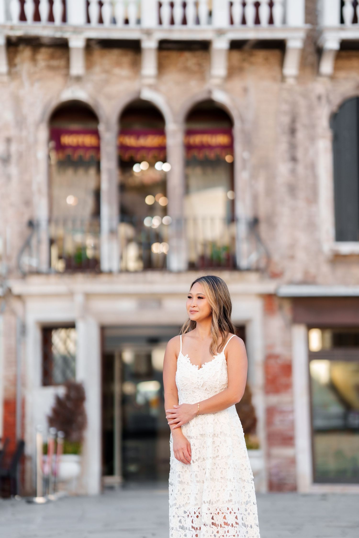 A woman in a white lace dress stands before a historic Venetian hotel building in Italy.