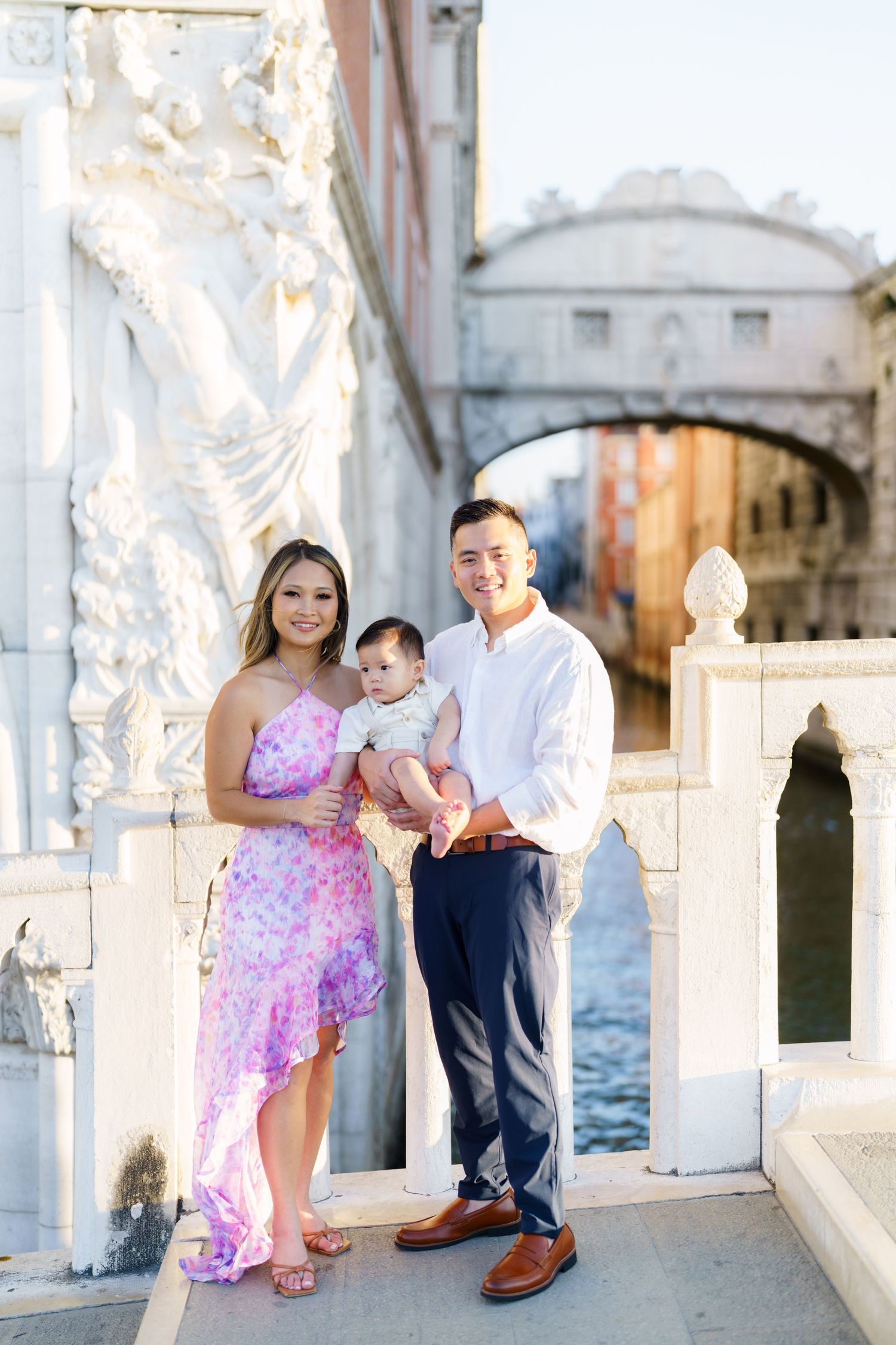 A young family poses with their baby on a Venice bridge, the Bridge of Sighs visible in the background.
