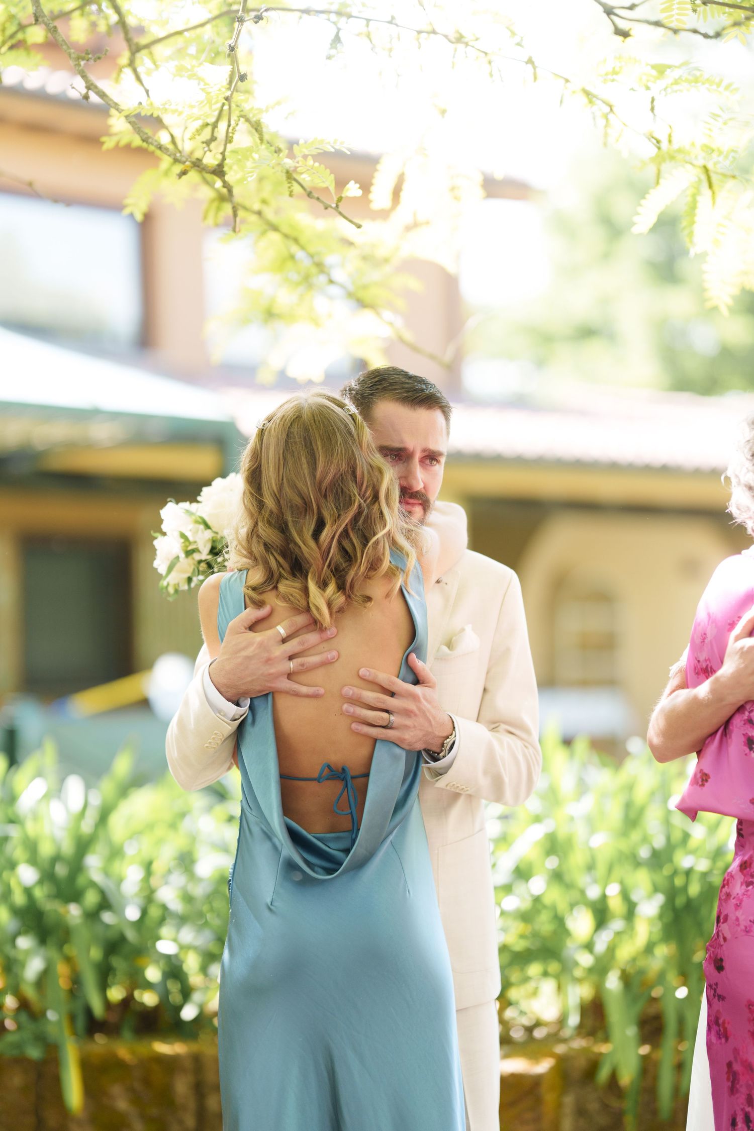 A man in a white suit embraces a woman in a blue backless dress outdoors at a garden wedding ceremony.