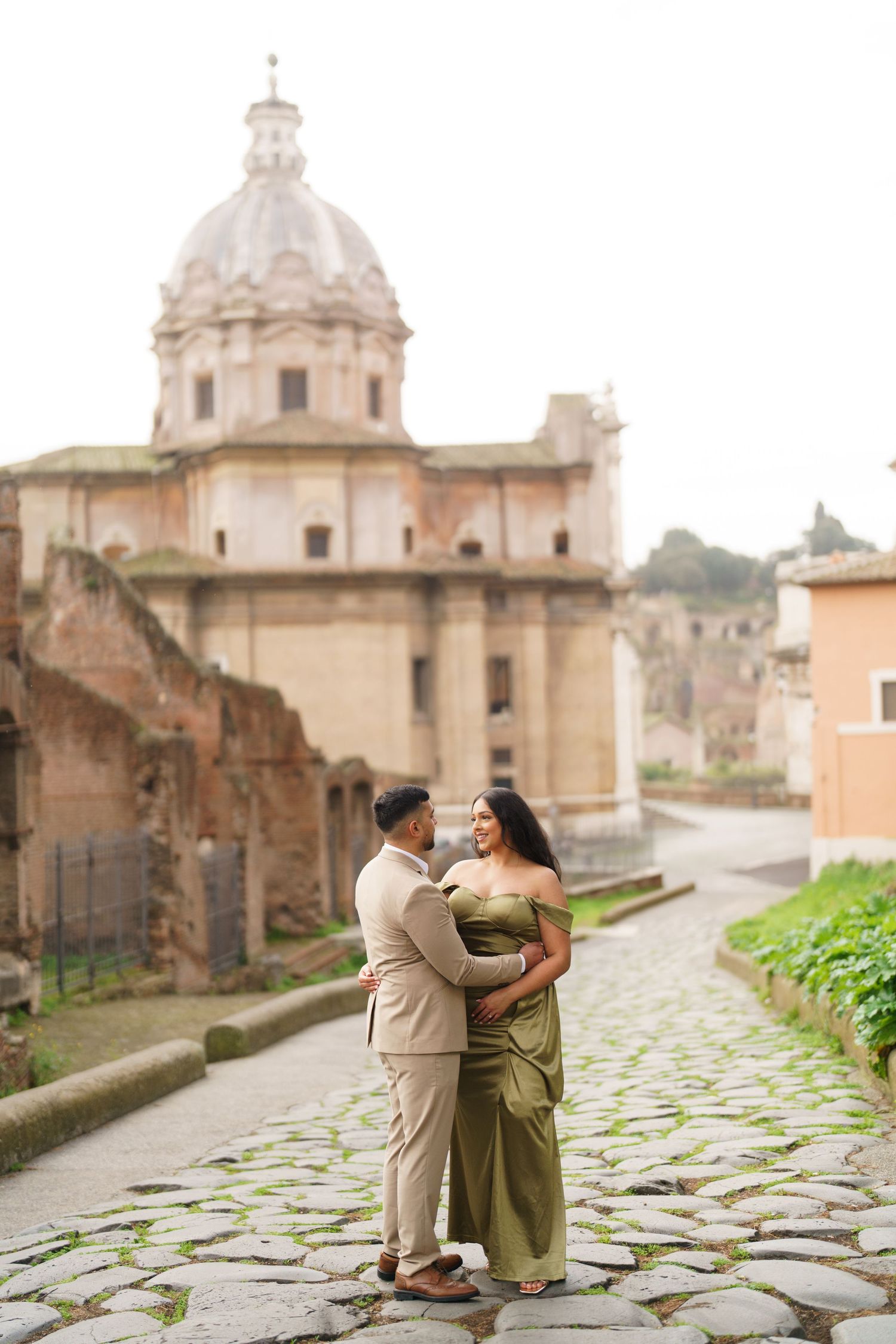 Couple embracing on cobblestone path in Rome, woman in green gown, man in tan suit, historic church dome backdrop.