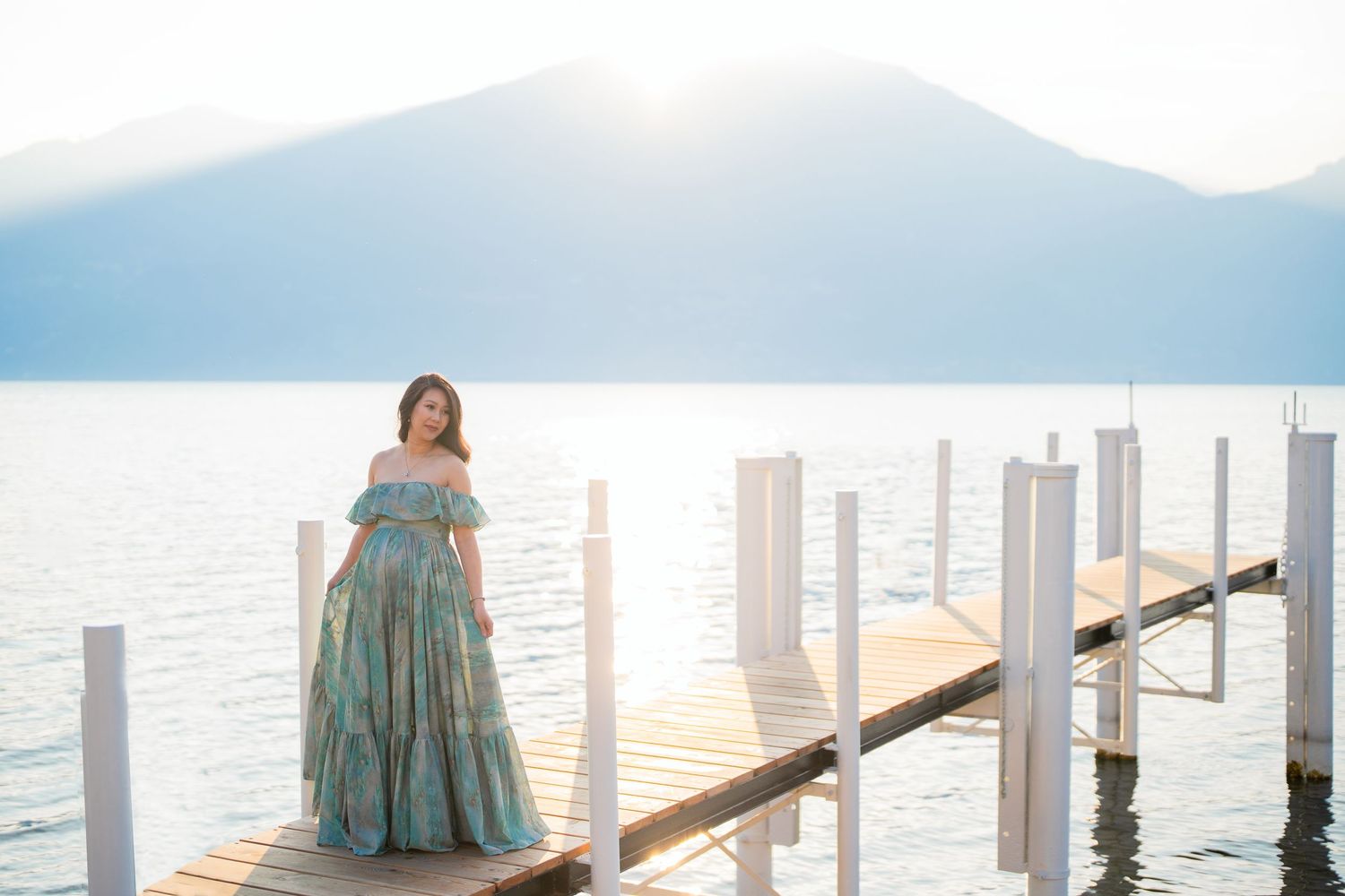 Pregnant woman in teal floral gown standing on wooden dock with mountain lake backdrop at sunset.