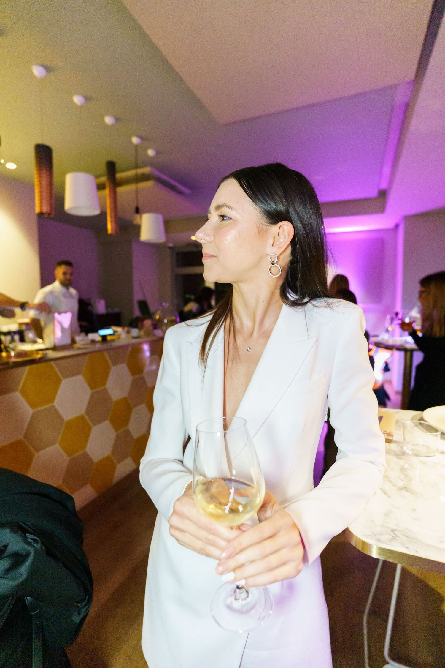 Elegant woman in white blazer holding wine glass at upscale bar with purple ambient lighting.