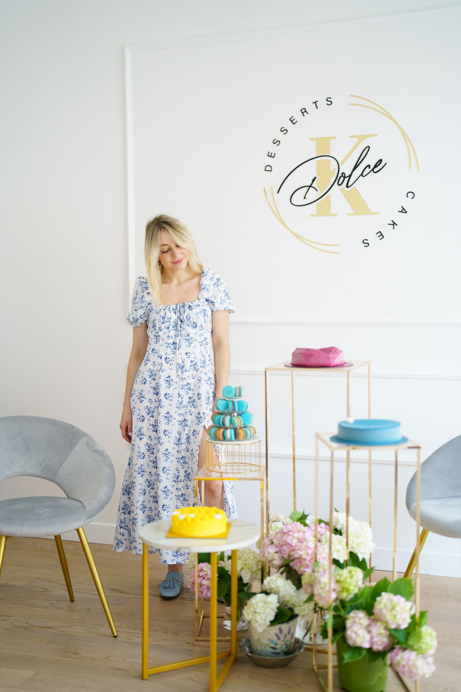 Blonde woman in floral dress stands beside elegant dessert display at Dolce K Cakes & Desserts boutique bakery.