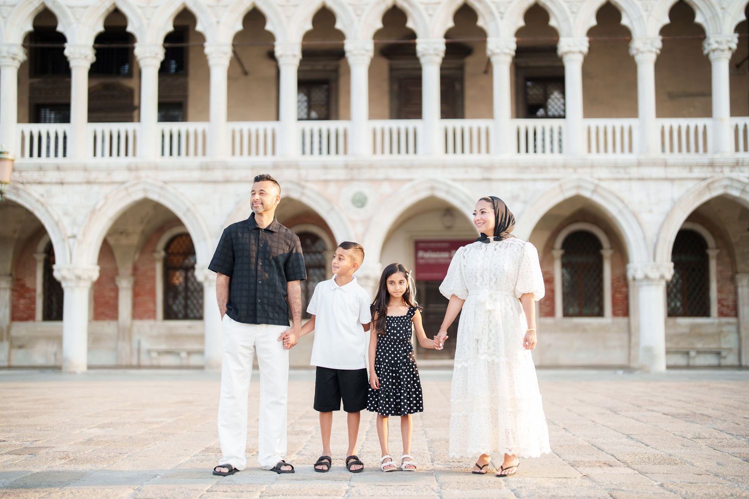 Family of four posing together in a grand Venetian arcade courtyard with ornate white arches.
