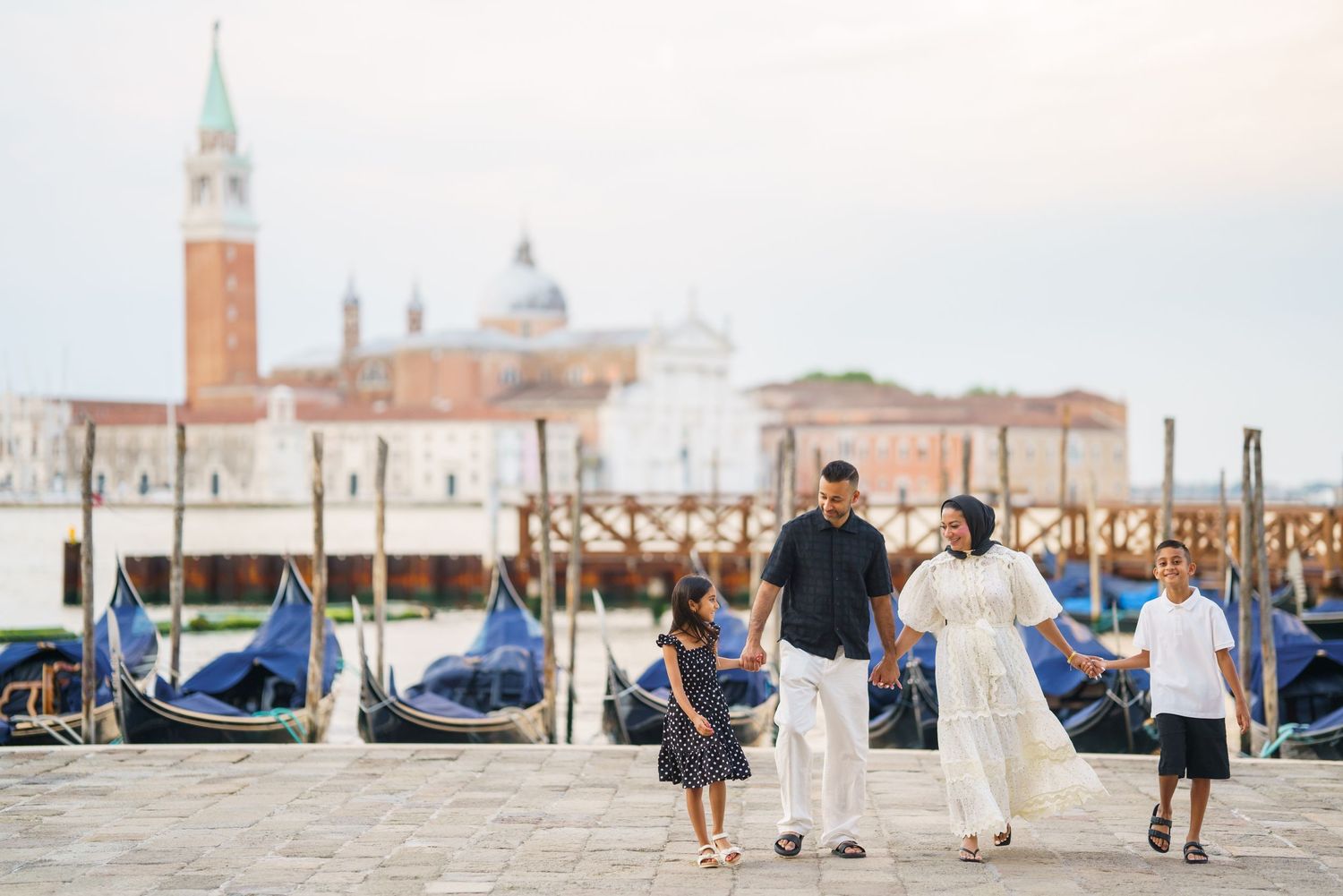 Family of four walking along Venice waterfront with gondolas and San Giorgio Maggiore church in background.