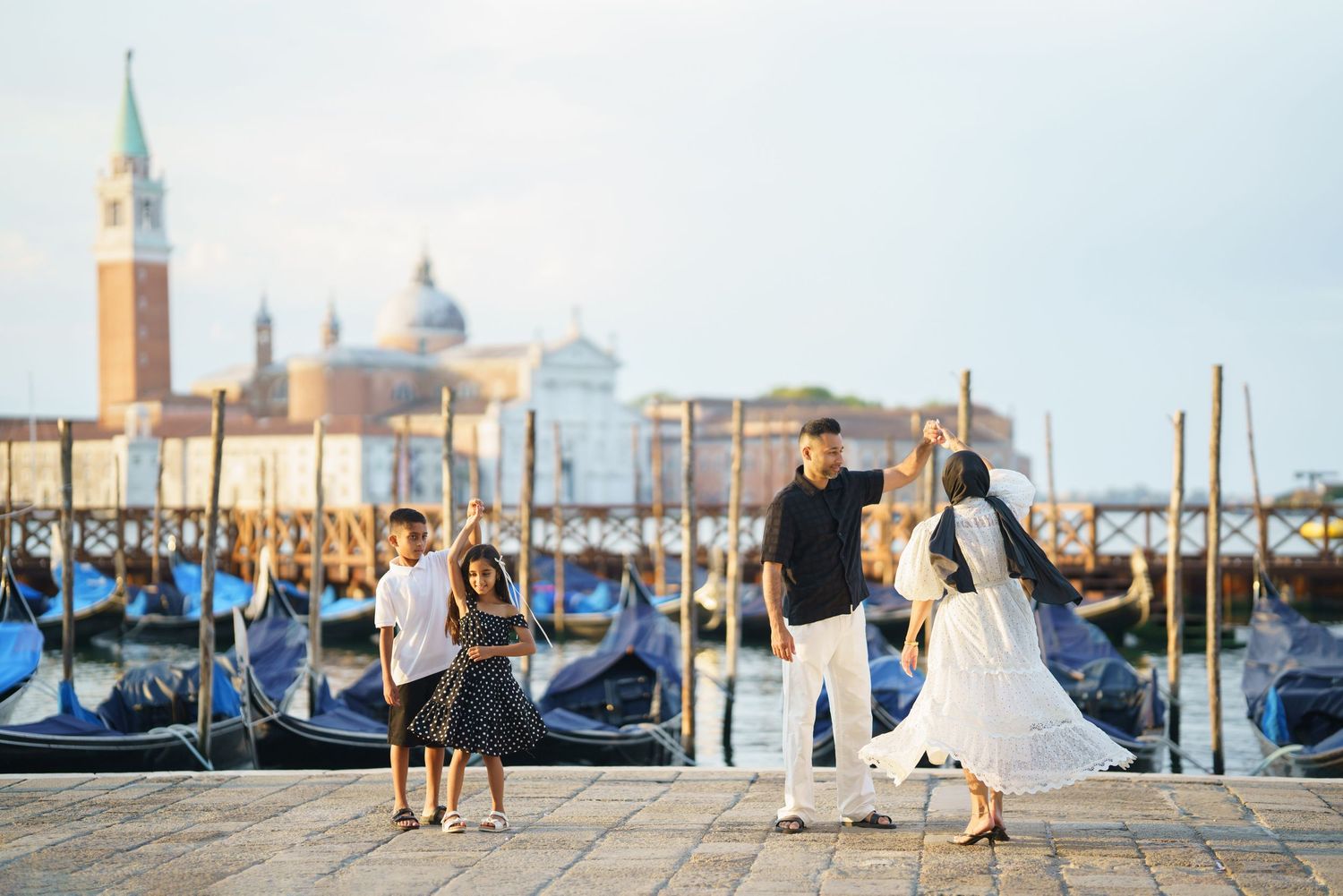 Family posing by gondolas on Venice waterfront with San Giorgio Maggiore church in background on sunny day.