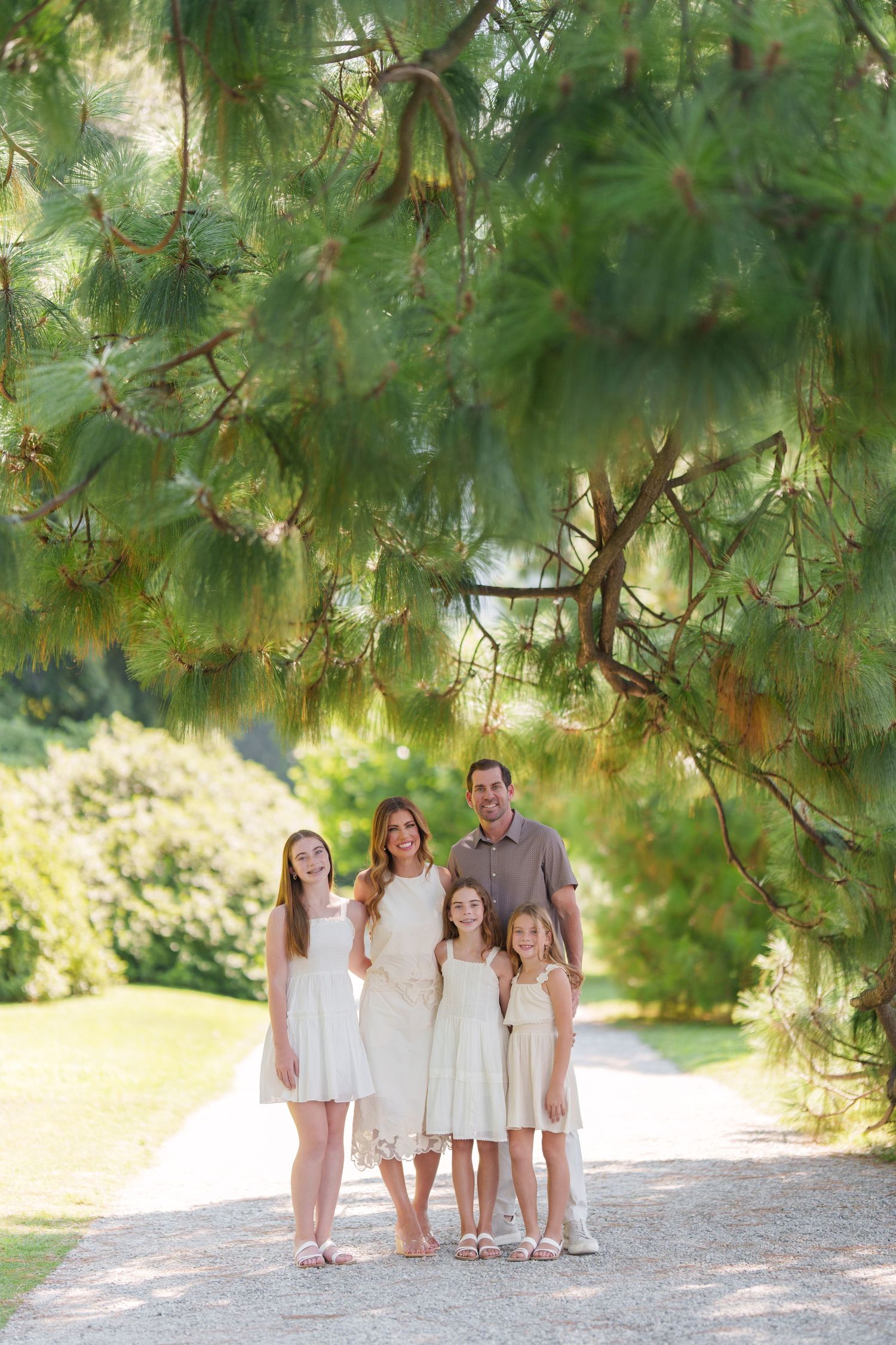 Family of five posing on a garden path under pine tree branches, wearing coordinated white and beige outfits.