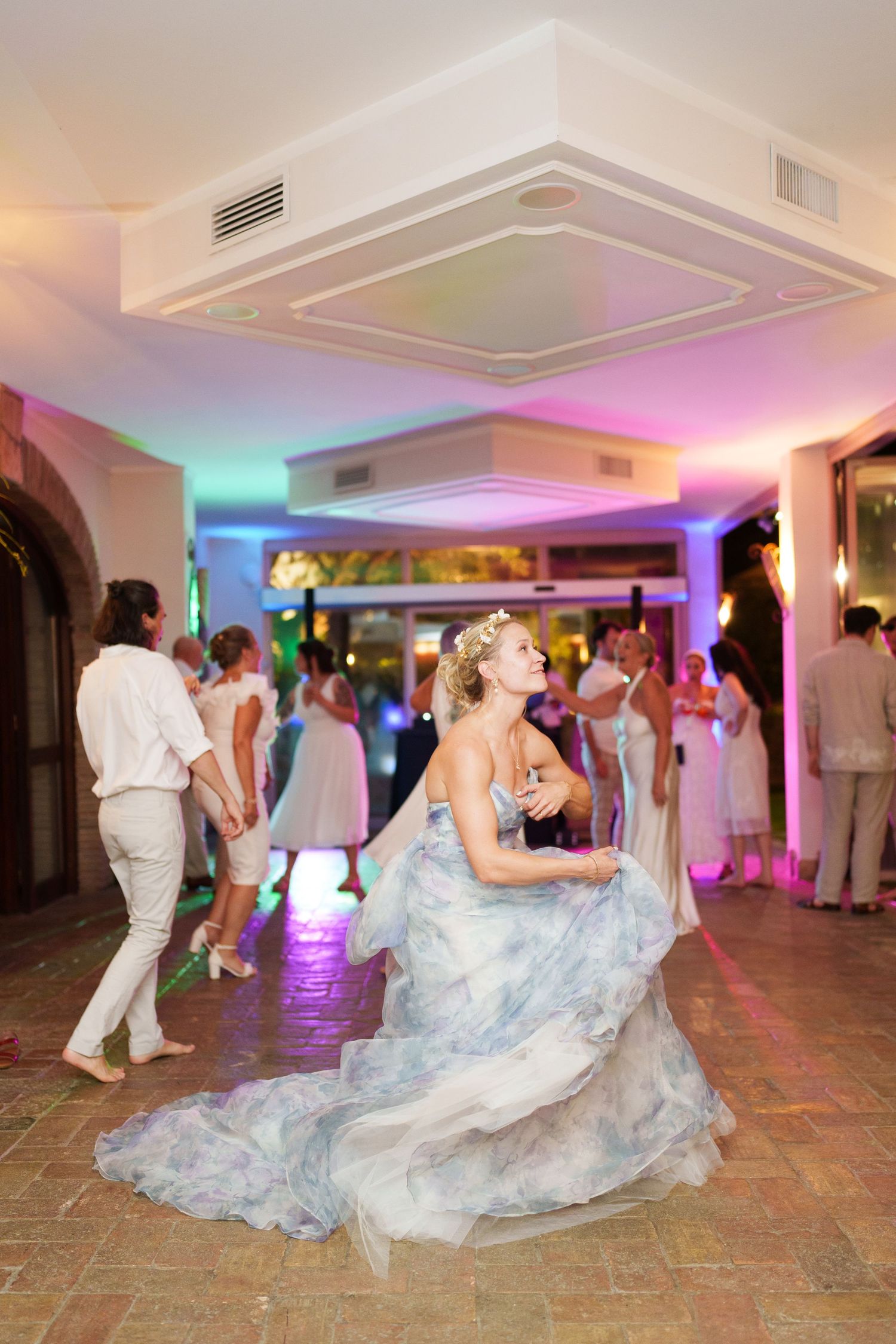 Bride in blue floral gown dancing joyfully at wedding reception with colorful lights and guests in background.