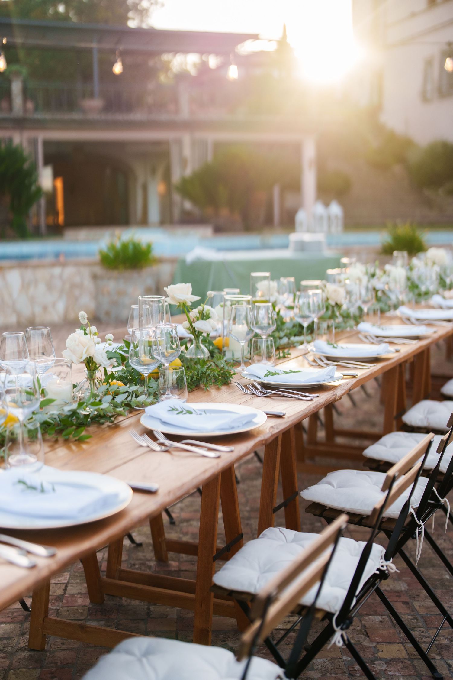 Elegant outdoor wedding reception table with floral garland, white plates, and glassware set at golden hour.