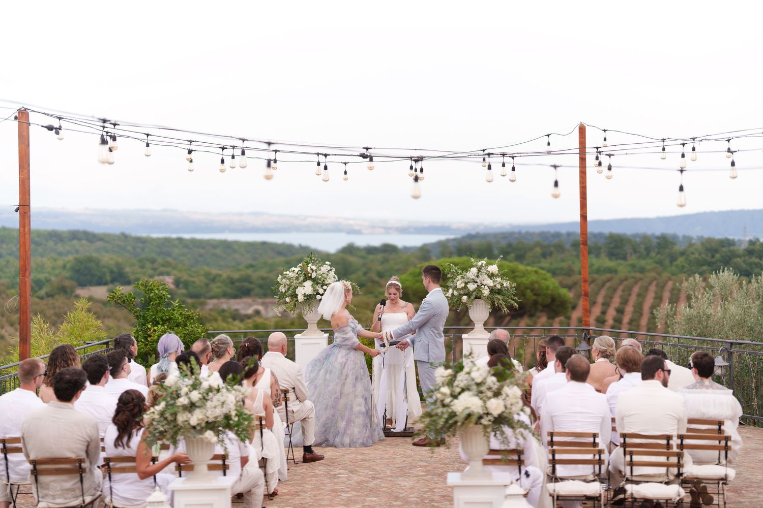 Outdoor wedding ceremony on a terrace with string lights, white floral arrangements, and a scenic vineyard valley view.