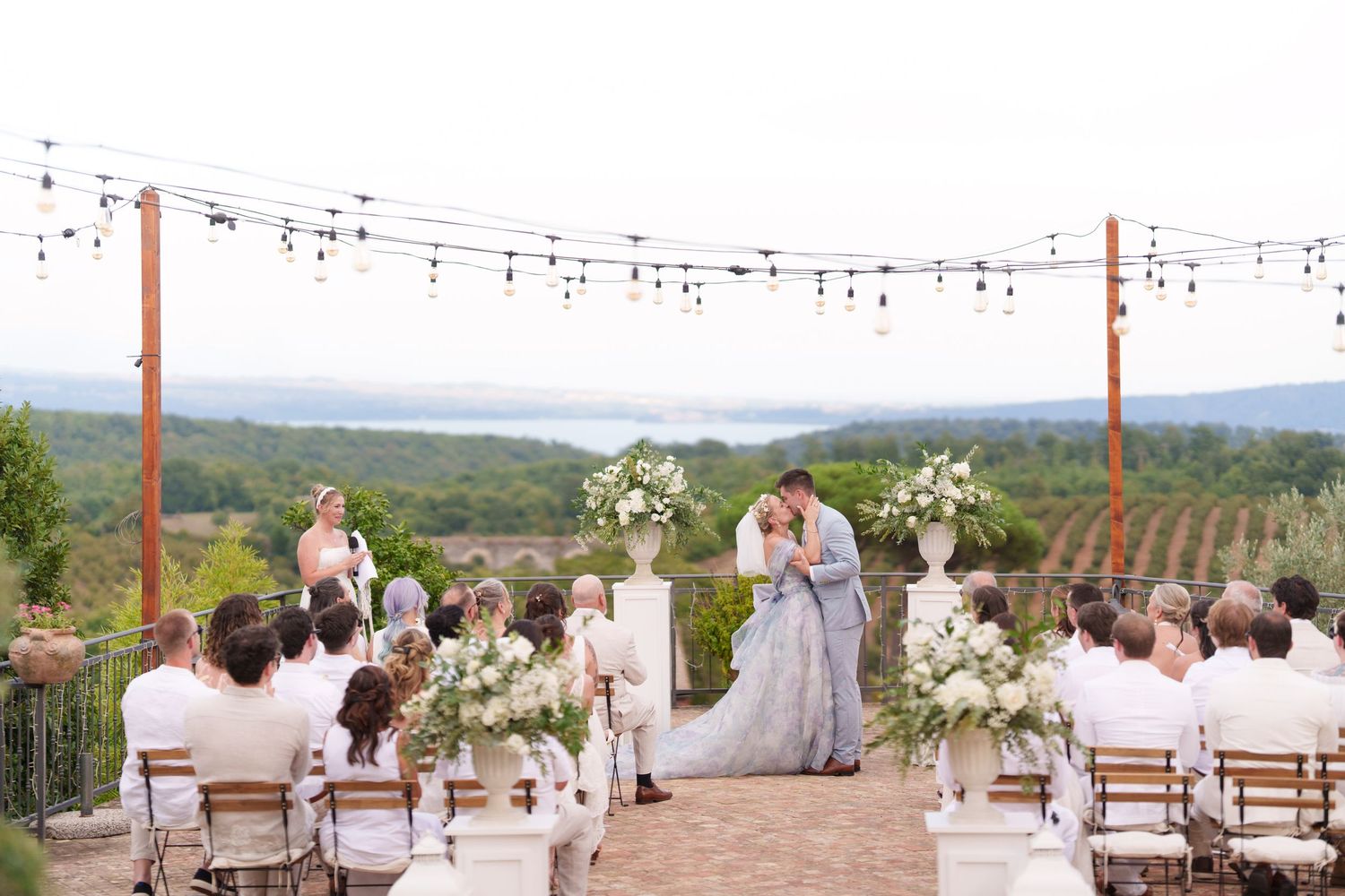 Outdoor vineyard wedding ceremony with couple at floral altar, string lights, and scenic lake views.