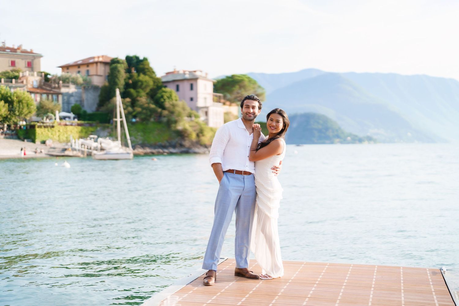 Couple in white outfits embracing on a dock with scenic lake and mountain views in the background.