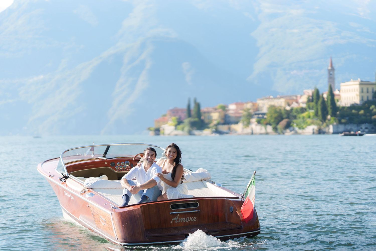 Couple riding a classic wooden boat on Lake Como, Italy, with scenic mountains and village in the background.