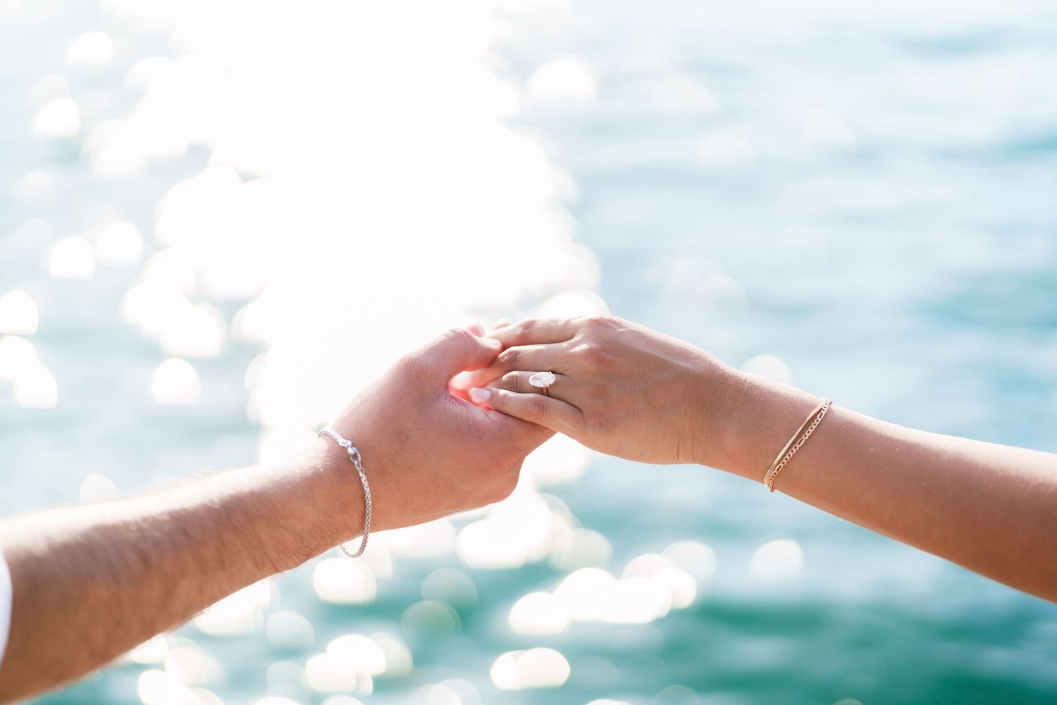 Two people gently holding hands with sparkling blue ocean water in the background on a sunny day.