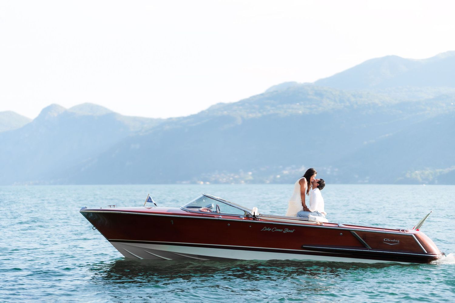A bride and groom share a romantic kiss on a classic wooden speedboat on a serene mountain lake.