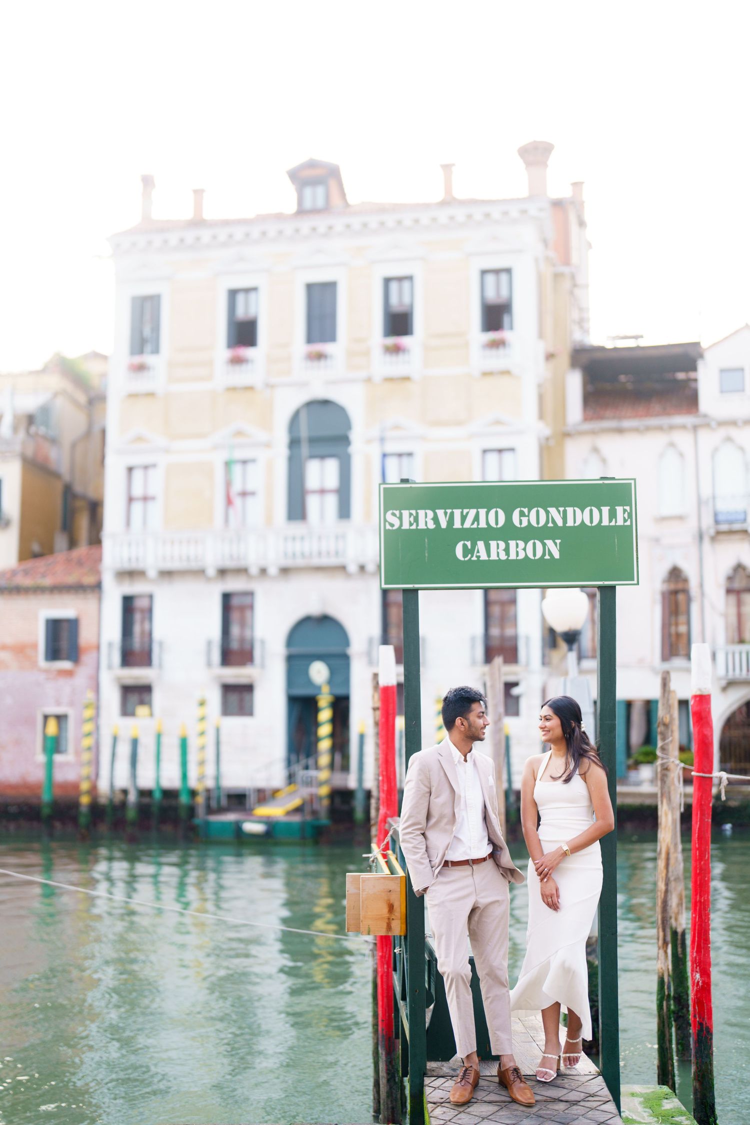 Couple in light-colored attire poses romantically by the iconic green gondola station posts along Venice's canal waterways.