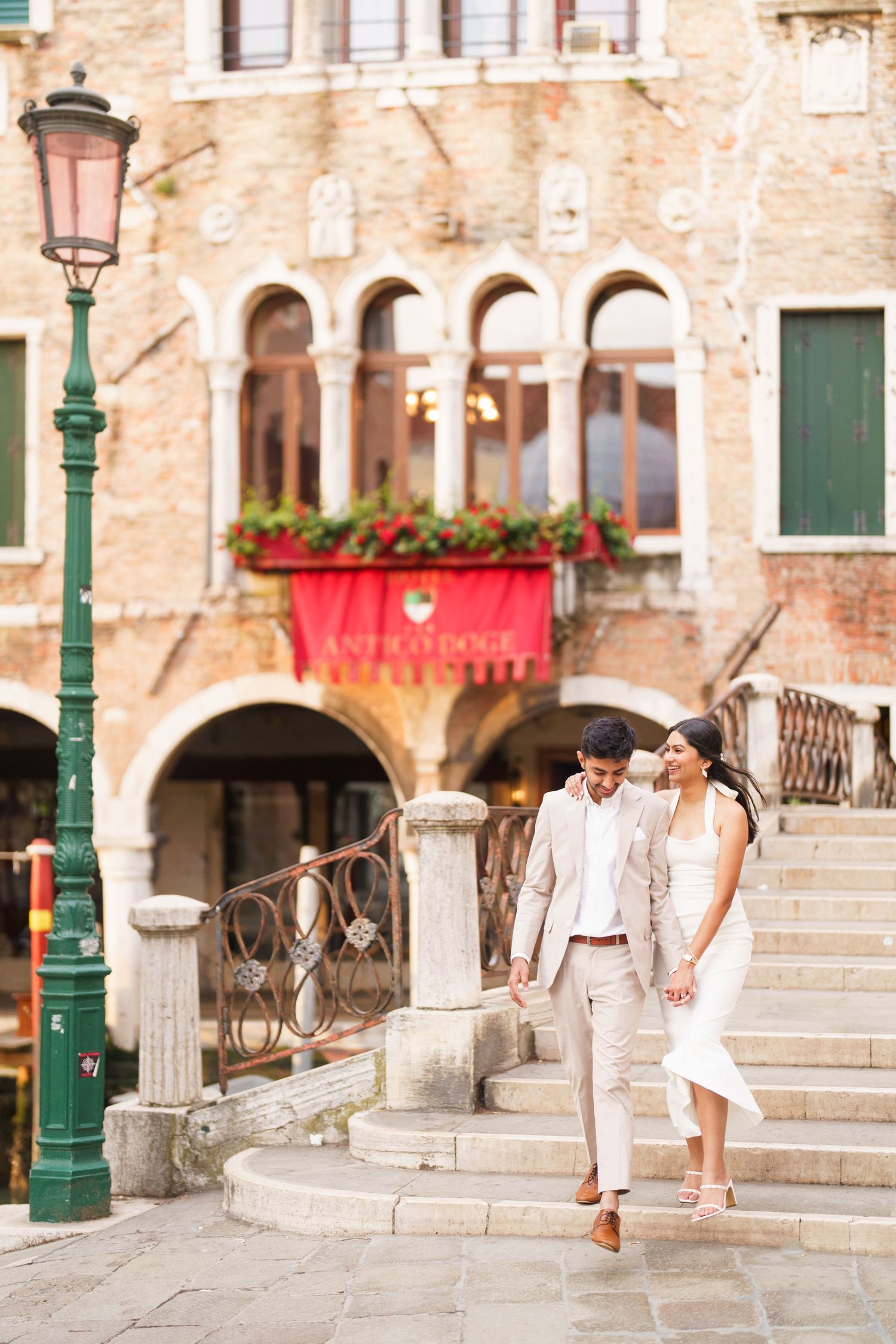 A couple in white outfits walks up stone steps in a romantic Venice setting with vintage lampposts and brick walls.