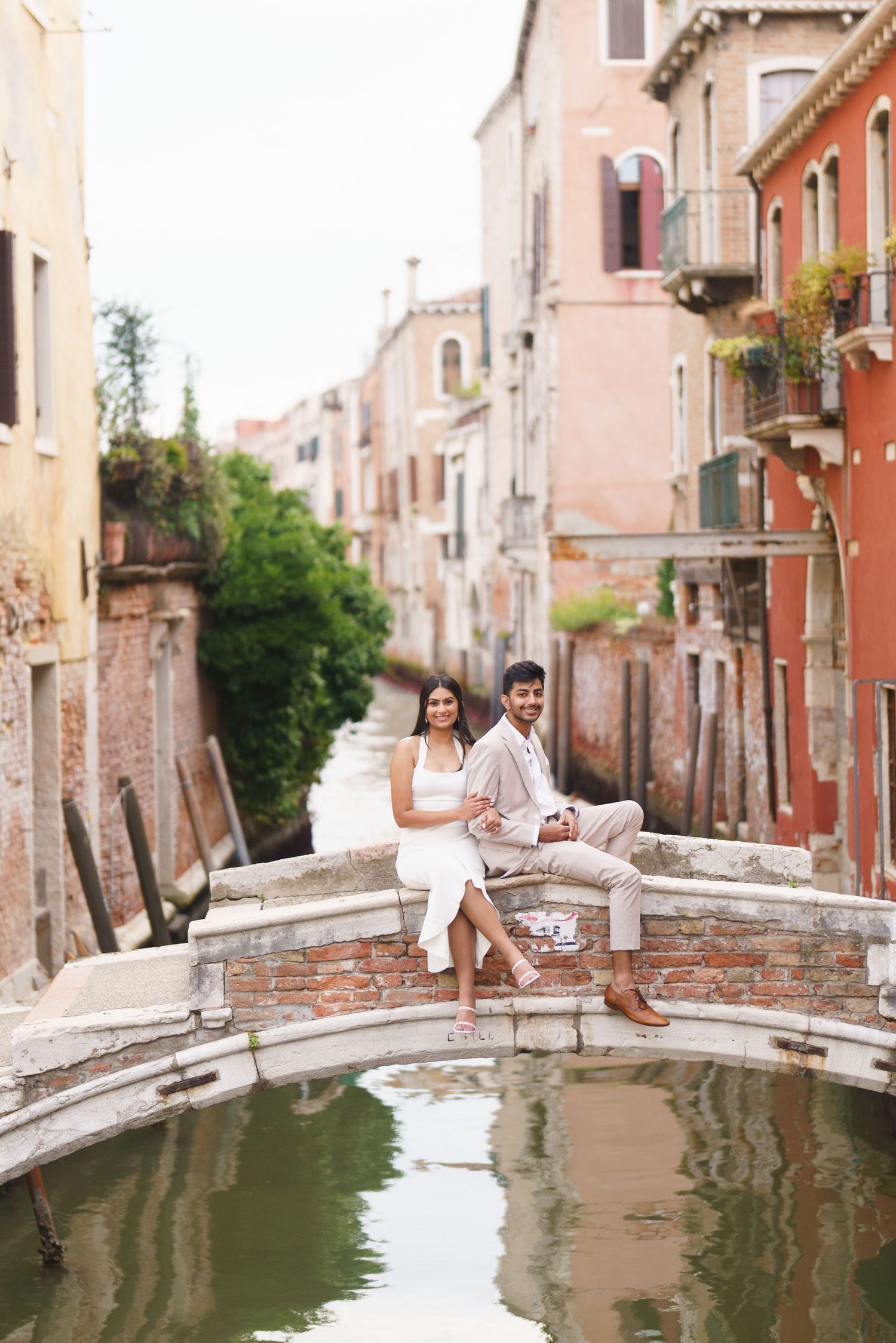 A couple sits together on a charming brick bridge over a canal in Venice, Italy with colorful buildings in the background.