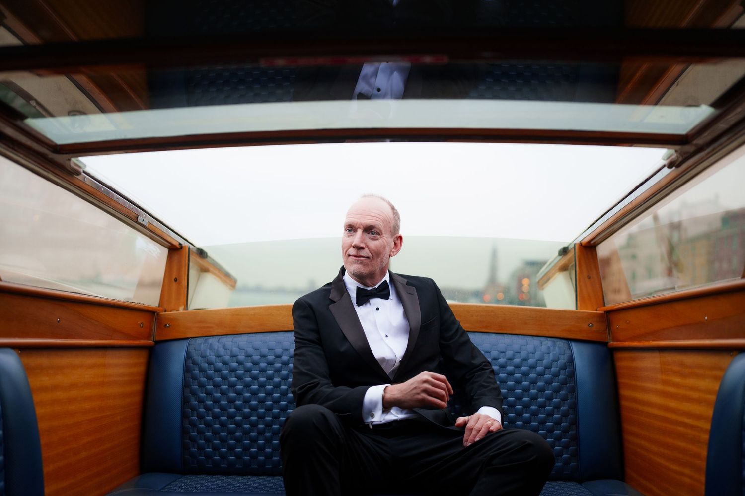 A person in a black tuxedo and bow tie sits on blue leather seating inside a luxury wooden boat cabin.