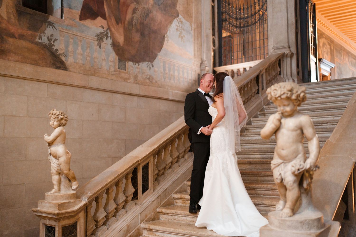 A bride and groom share an intimate moment on an elegant marble staircase adorned with classical statues.