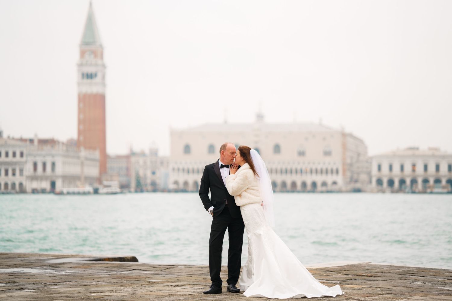 Romantic wedding couple embraces along the Venice waterfront with historic buildings and canals in the background.