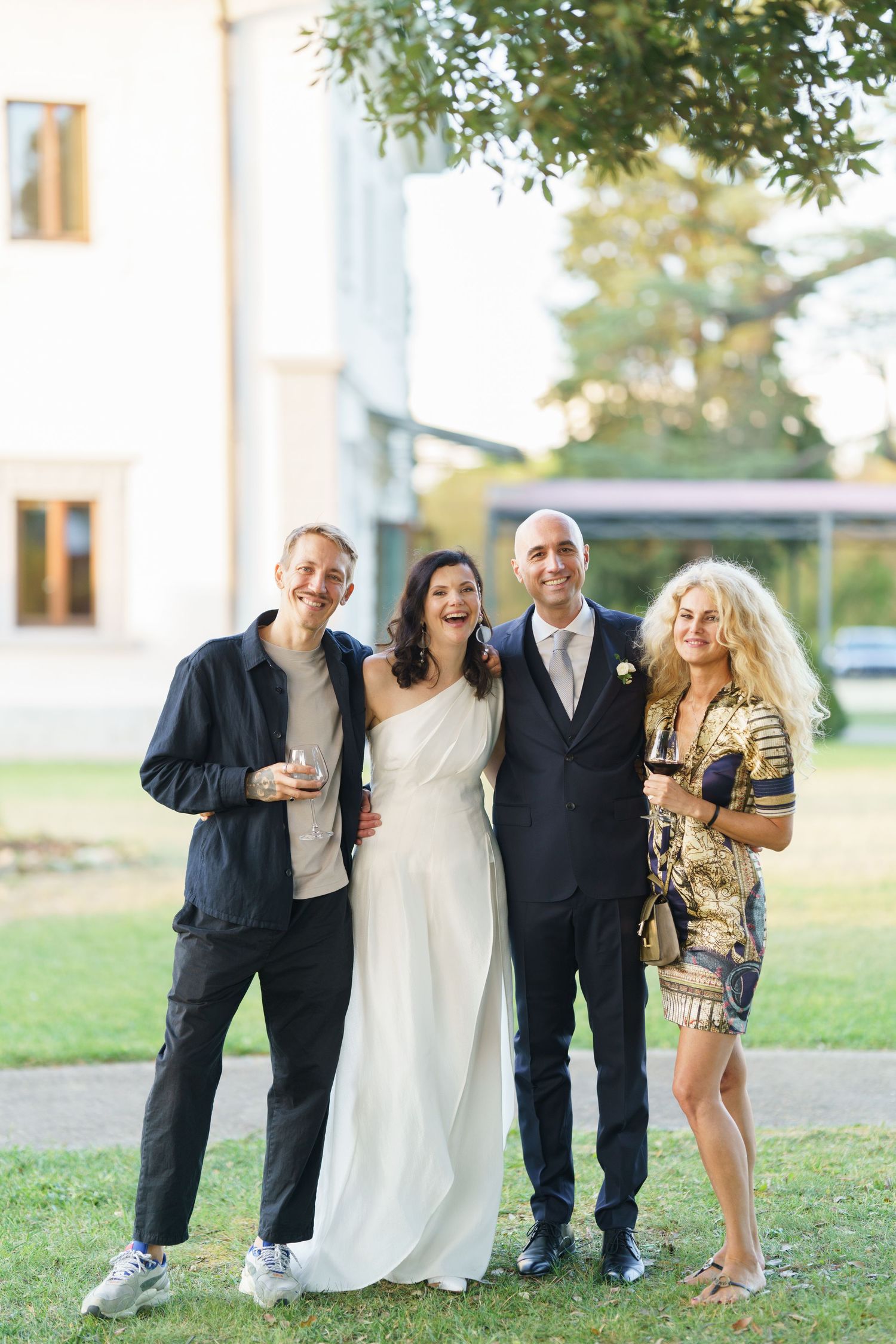 Wedding group poses playfully on grass in front of white building.