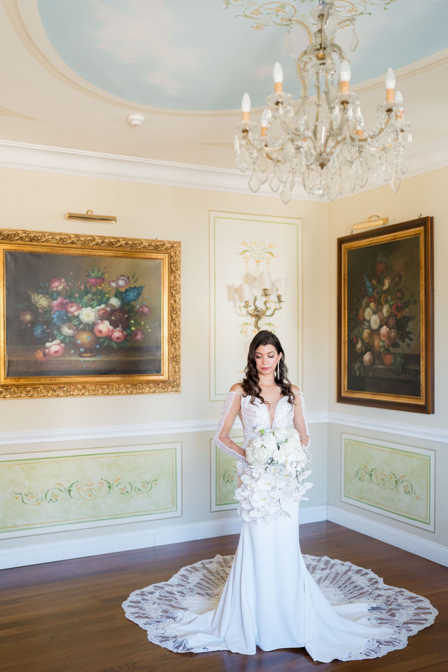 Bride in white gown poses in elegant room with crystal chandelier and classic artwork.