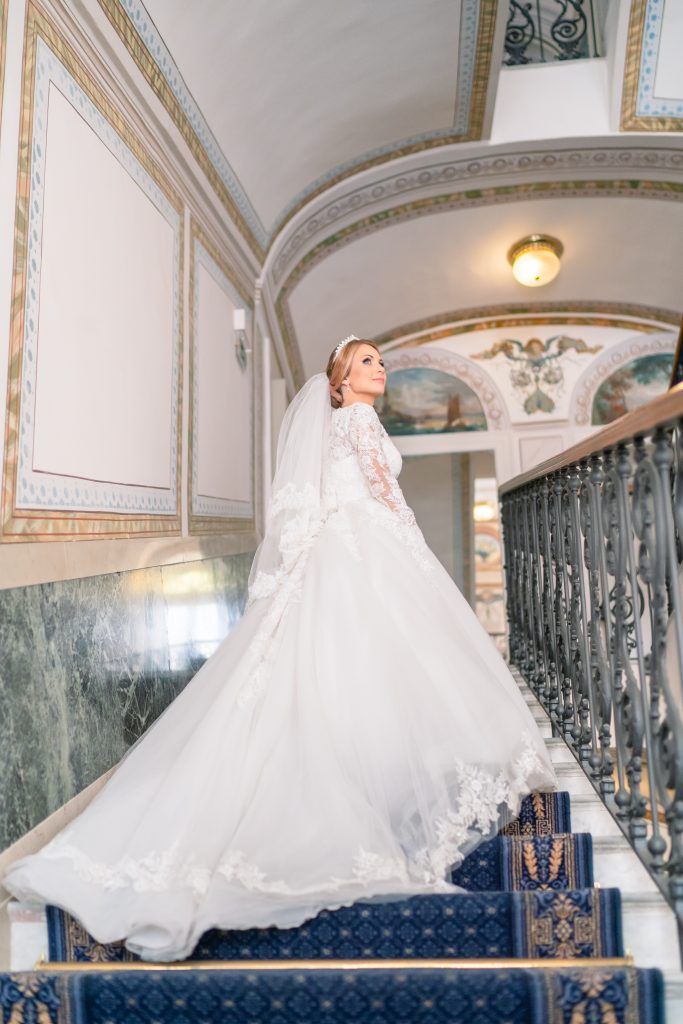 Bride in flowing ballgown twirling on elegant staircase with decorative marble walls.