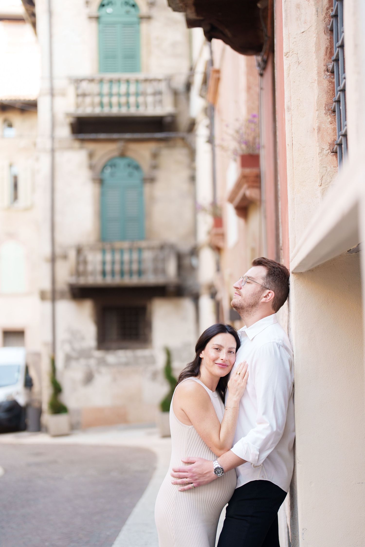 A couple embraces against a wall in a historic European street with turquoise shuttered windows and balconies.