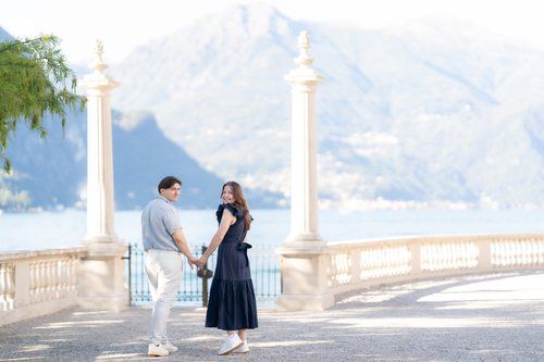 A couple shares a romantic moment on a white stone balcony overlooking mountains and water on a sunny day.
