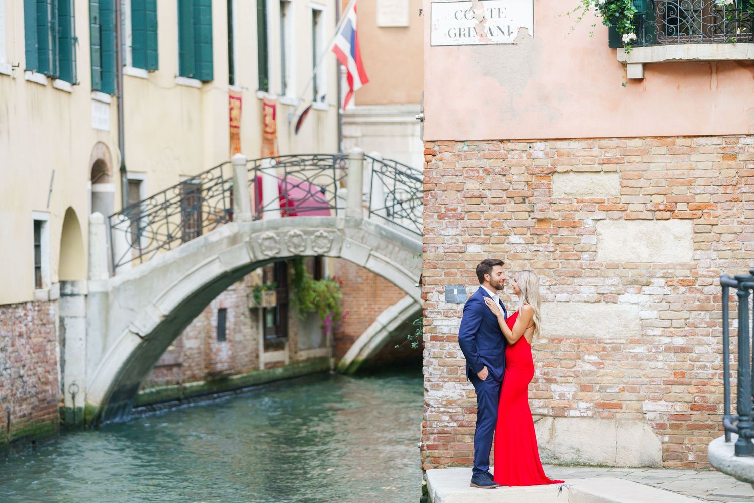 A couple in formal attire stands by a brick wall near a scenic Venice canal with an arched bridge in the background.