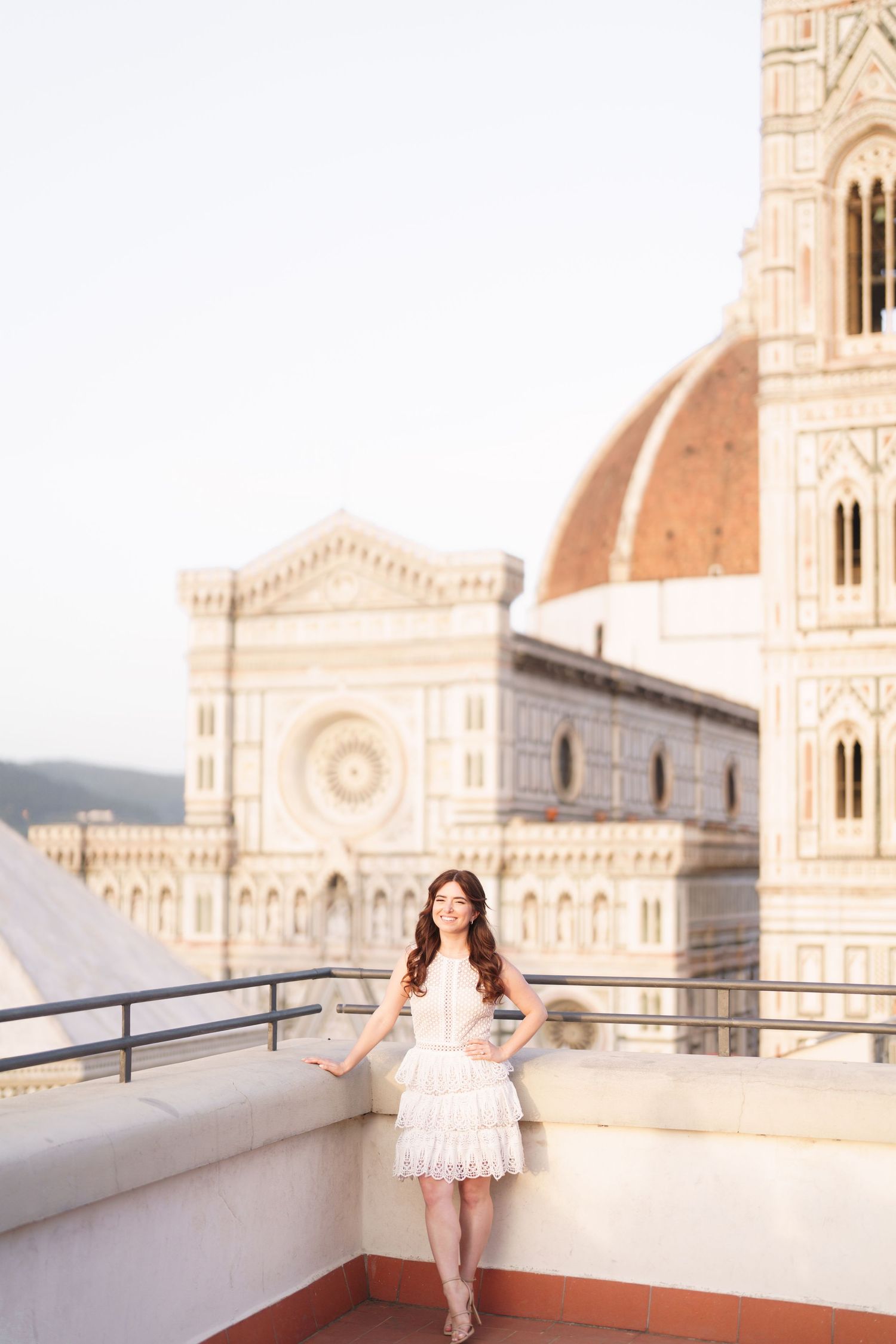 A woman in a white lace dress poses on a rooftop terrace overlooking the Florence Cathedral and Duomo in Italy.