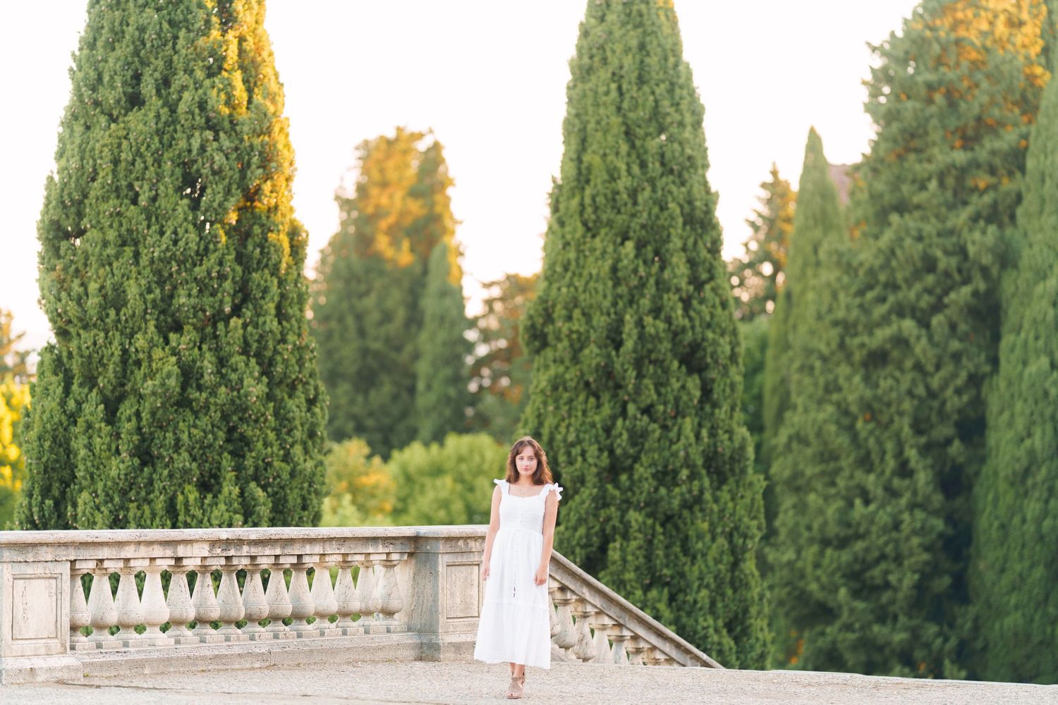 Someone in a white dress stands between tall cypress trees on a stone terrace at golden hour.