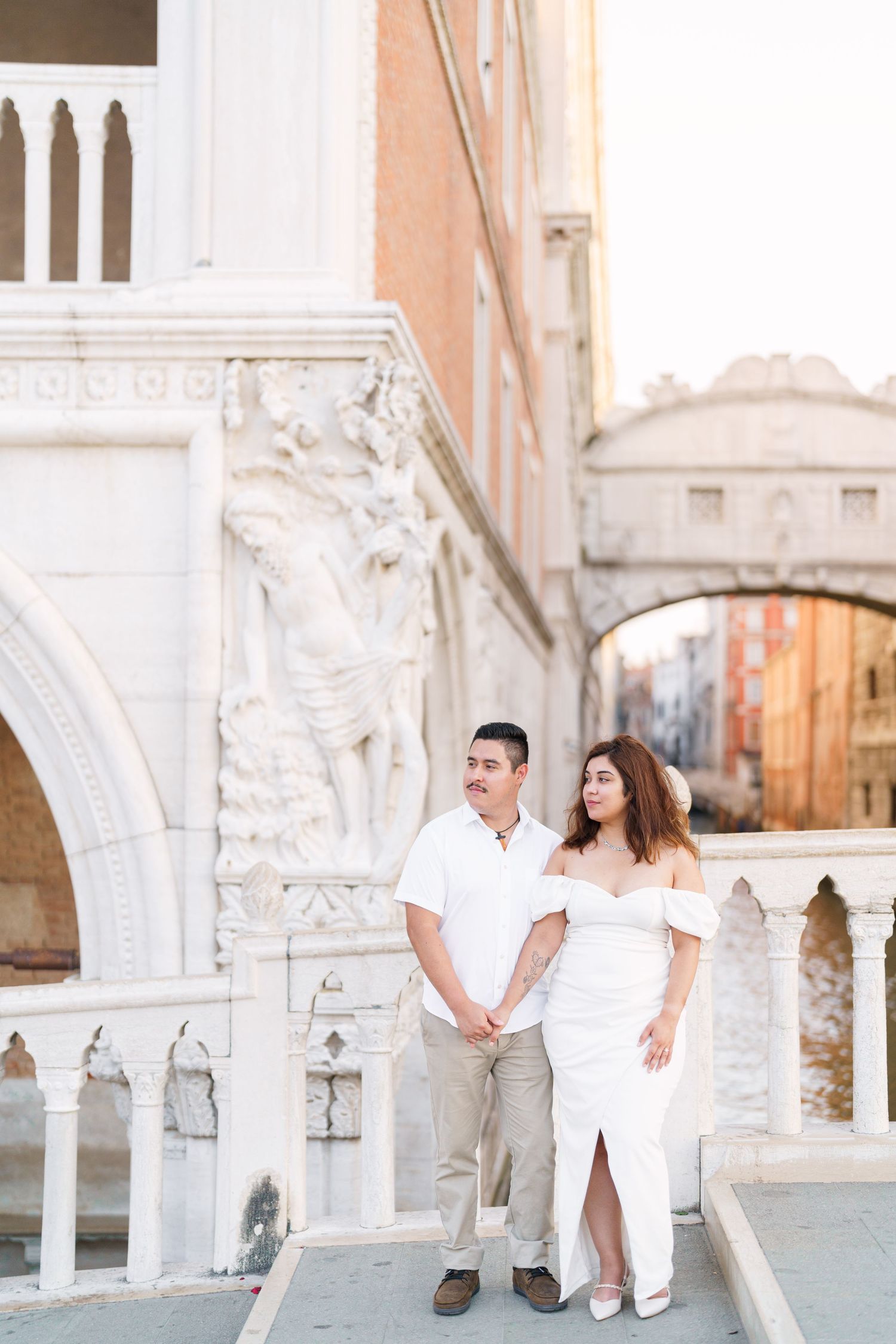 A couple poses together on a bridge in Venice, Italy with ornate white marble architecture and arches in the background.