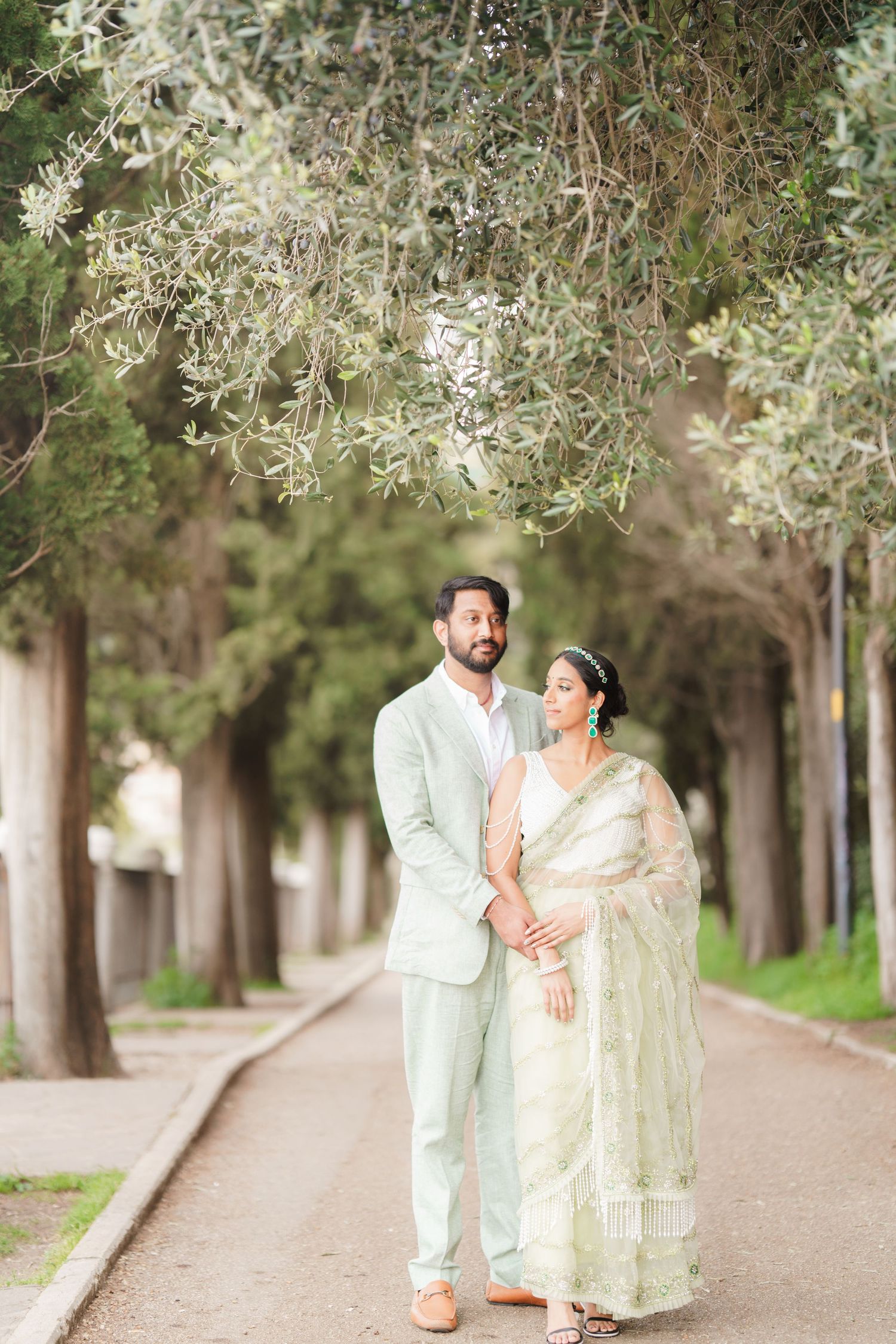 A couple in traditional white attire walks along a tree-lined path during a romantic outdoor photoshoot.