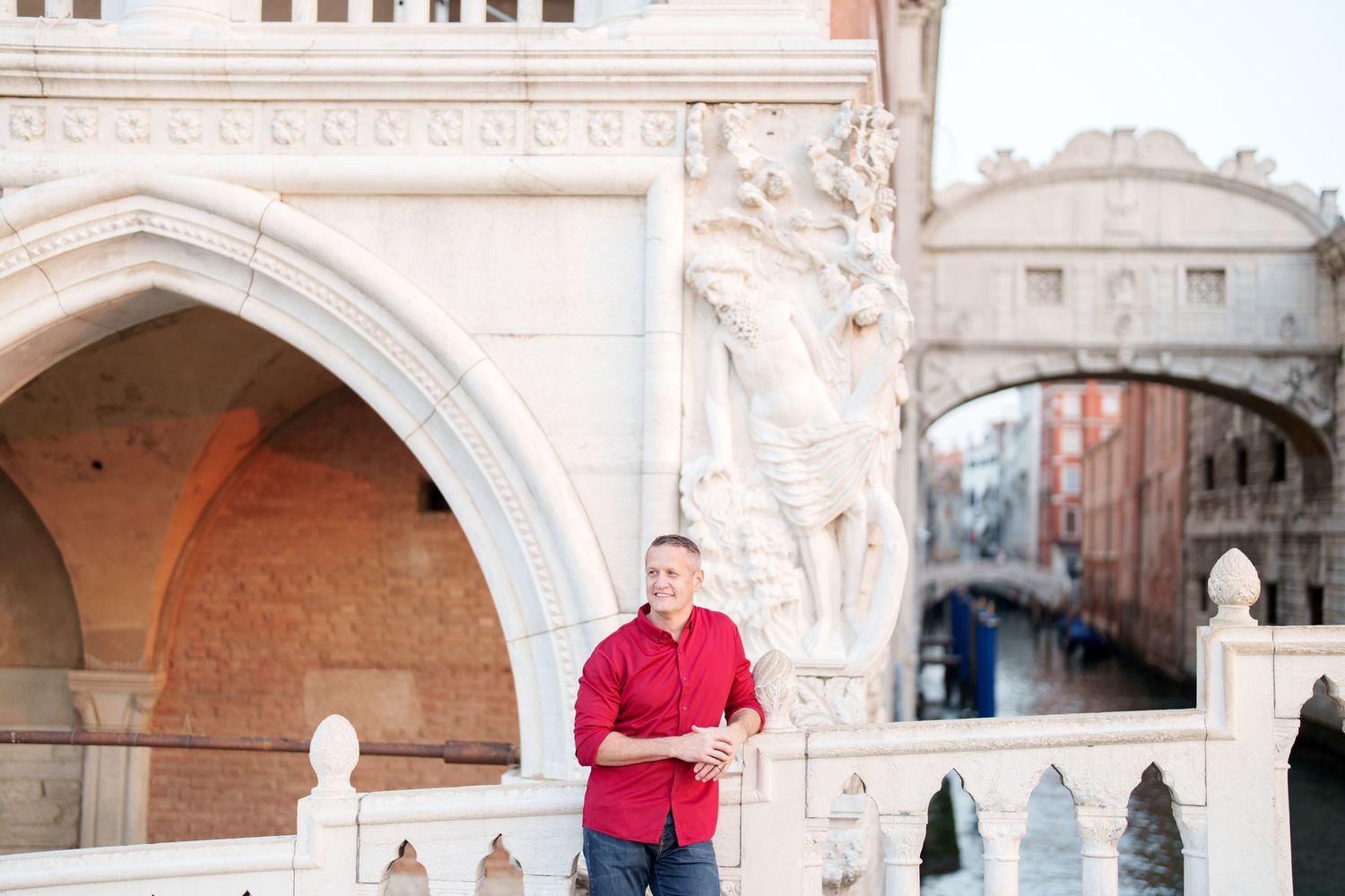 Person in red shirt stands beneath ornate stone archways at a historic European architectural site.